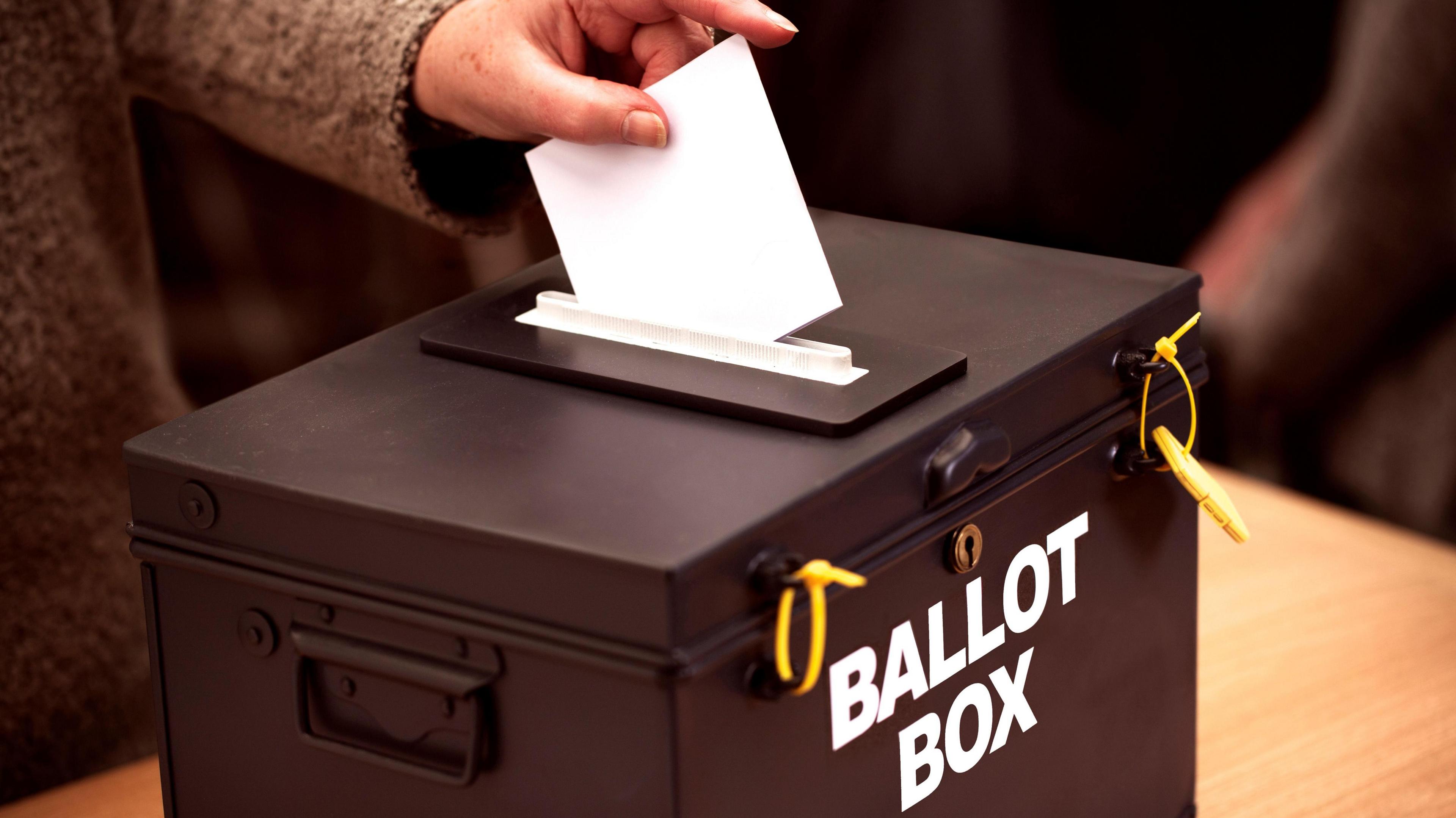 A close up of a hand putting a white paper voting slip into a black ballot box