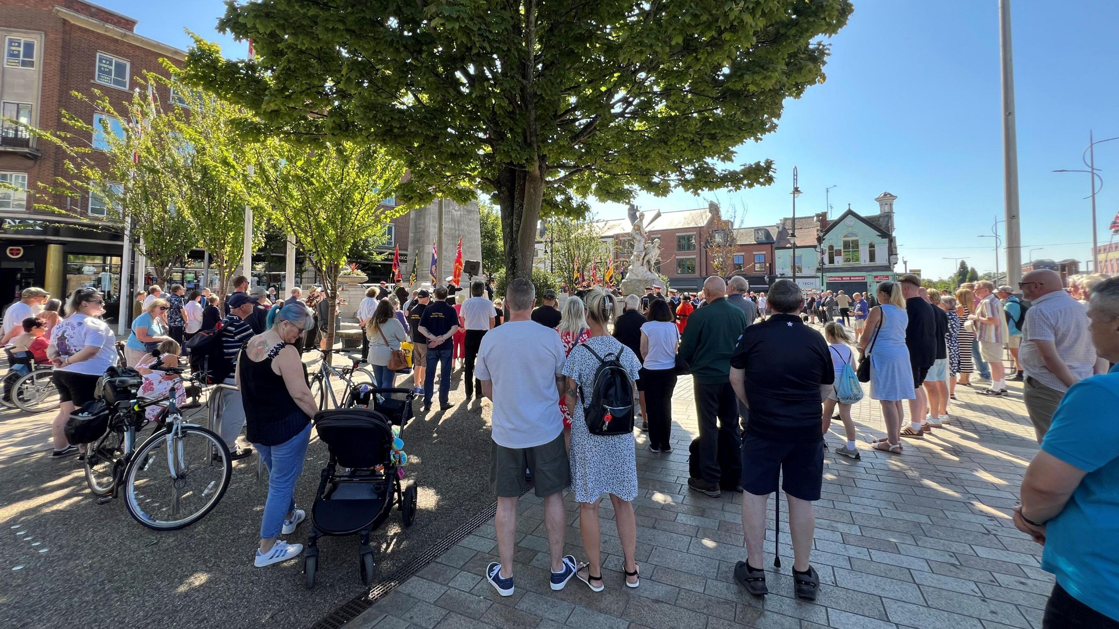 A crowd of people gathered on a paved square, with a large tree in the centre. The people are facing away from the camera and towards a ceremony. In the background are shops and other buildings. The sky is blue.