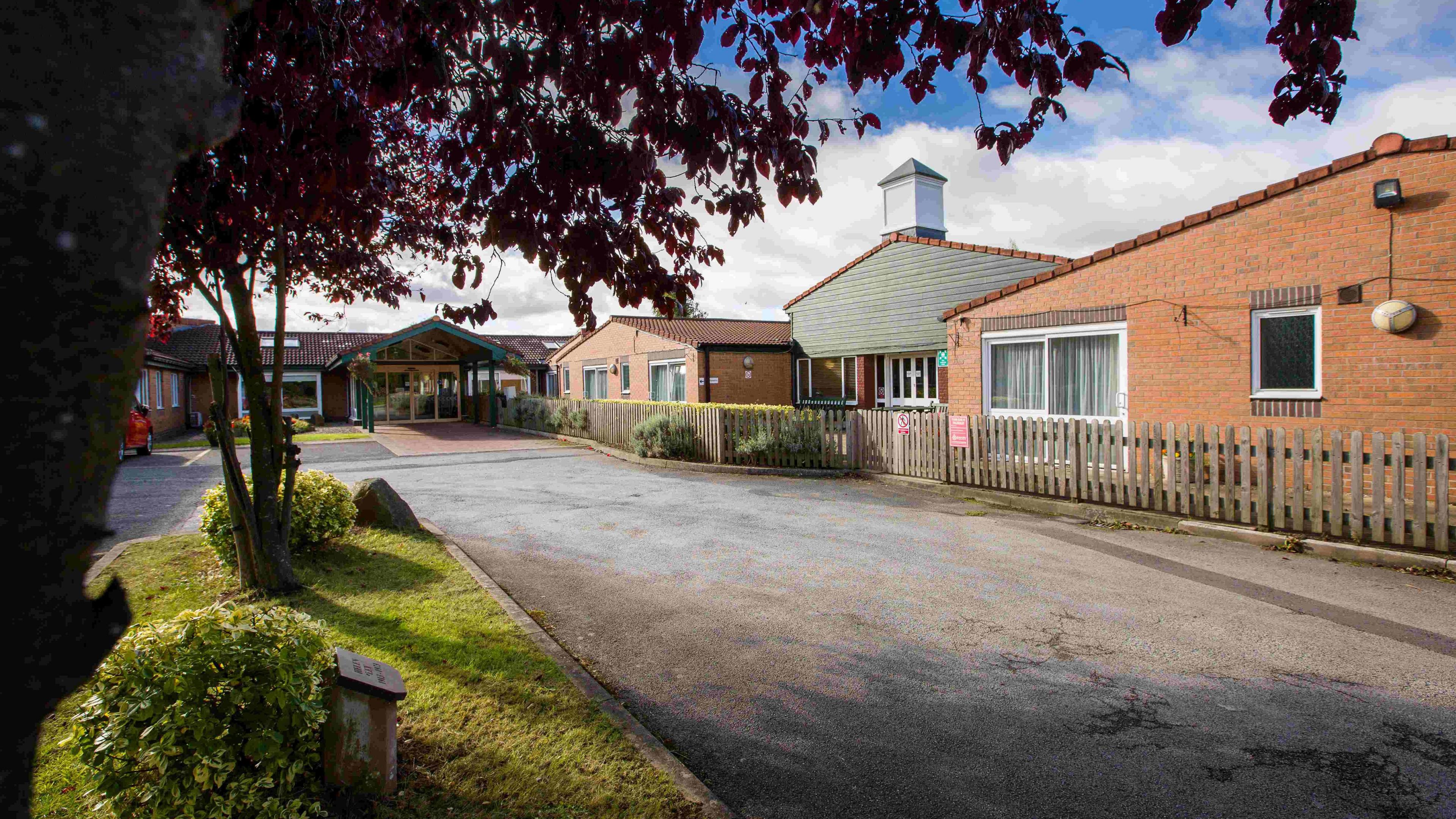 A single  storey red-brick building comprising of a number of joined pitched-roof structures. There is a long drive and car parking in front of the building.