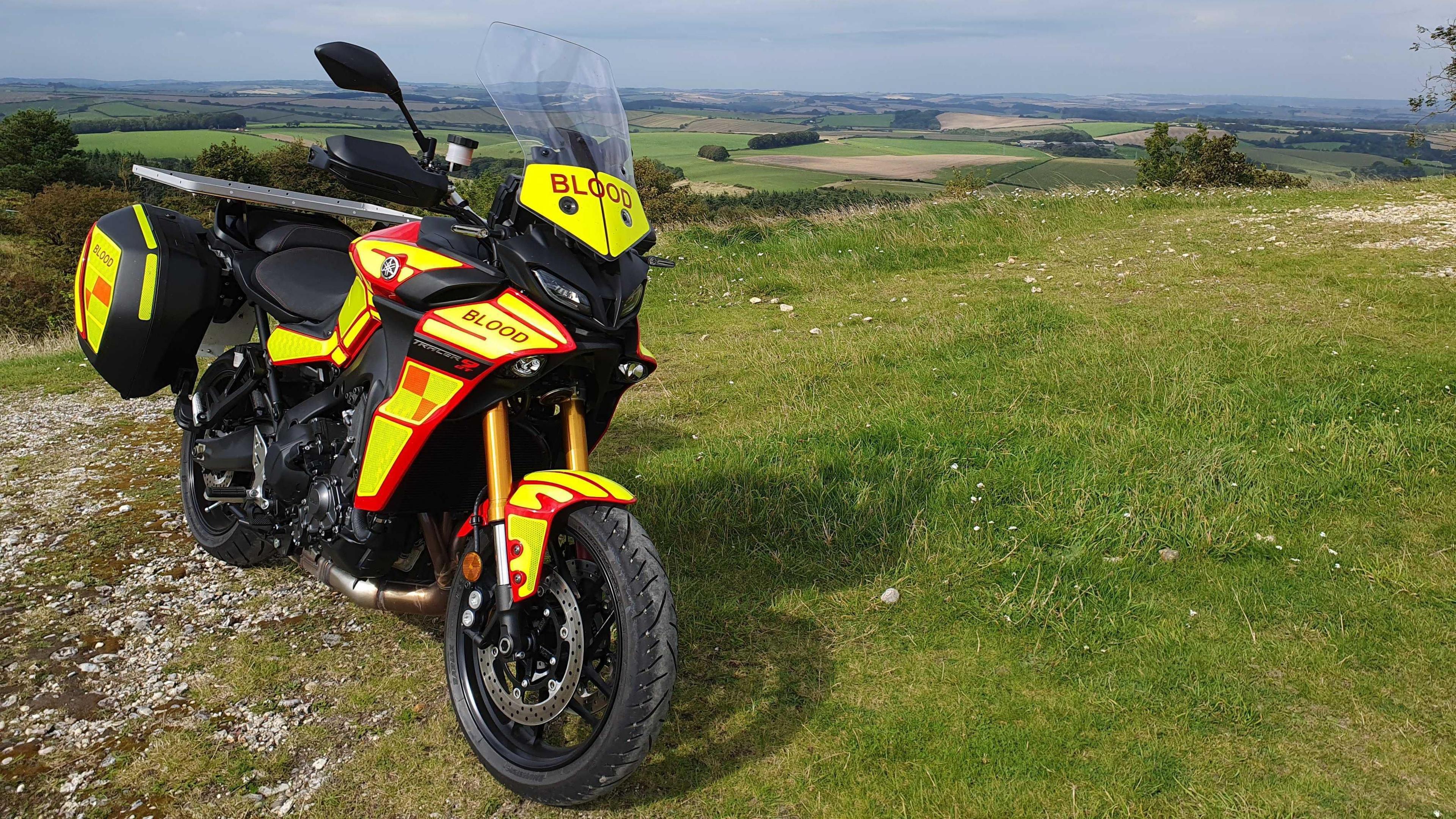 An emergency blood motorbike parked on a grassy hill overlooking fields stretching into the distance. It is a black motorbike with yellow and red hi-vis vinyl covering it, and the word 'Blood' written on every side. 