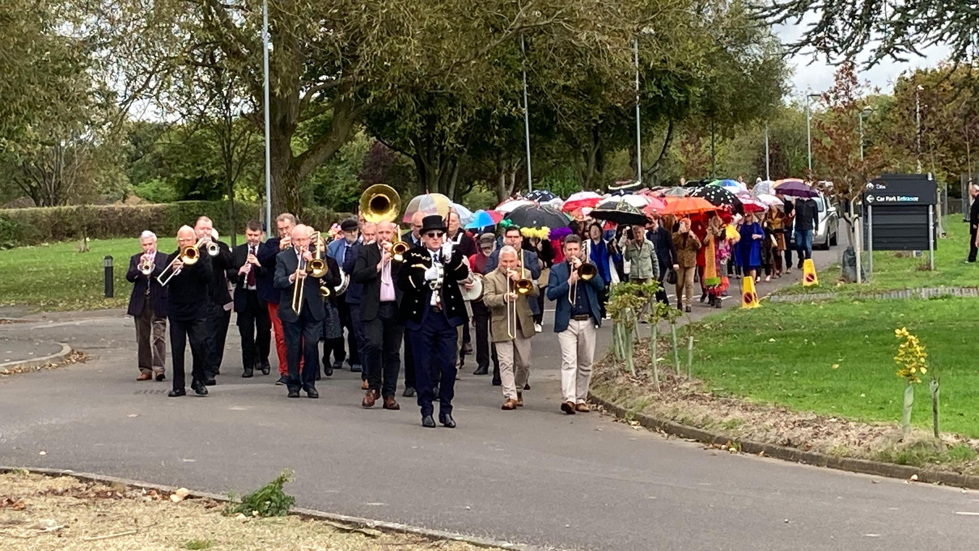 A funeral procession including a marching band and people holding up colourful umbrellas