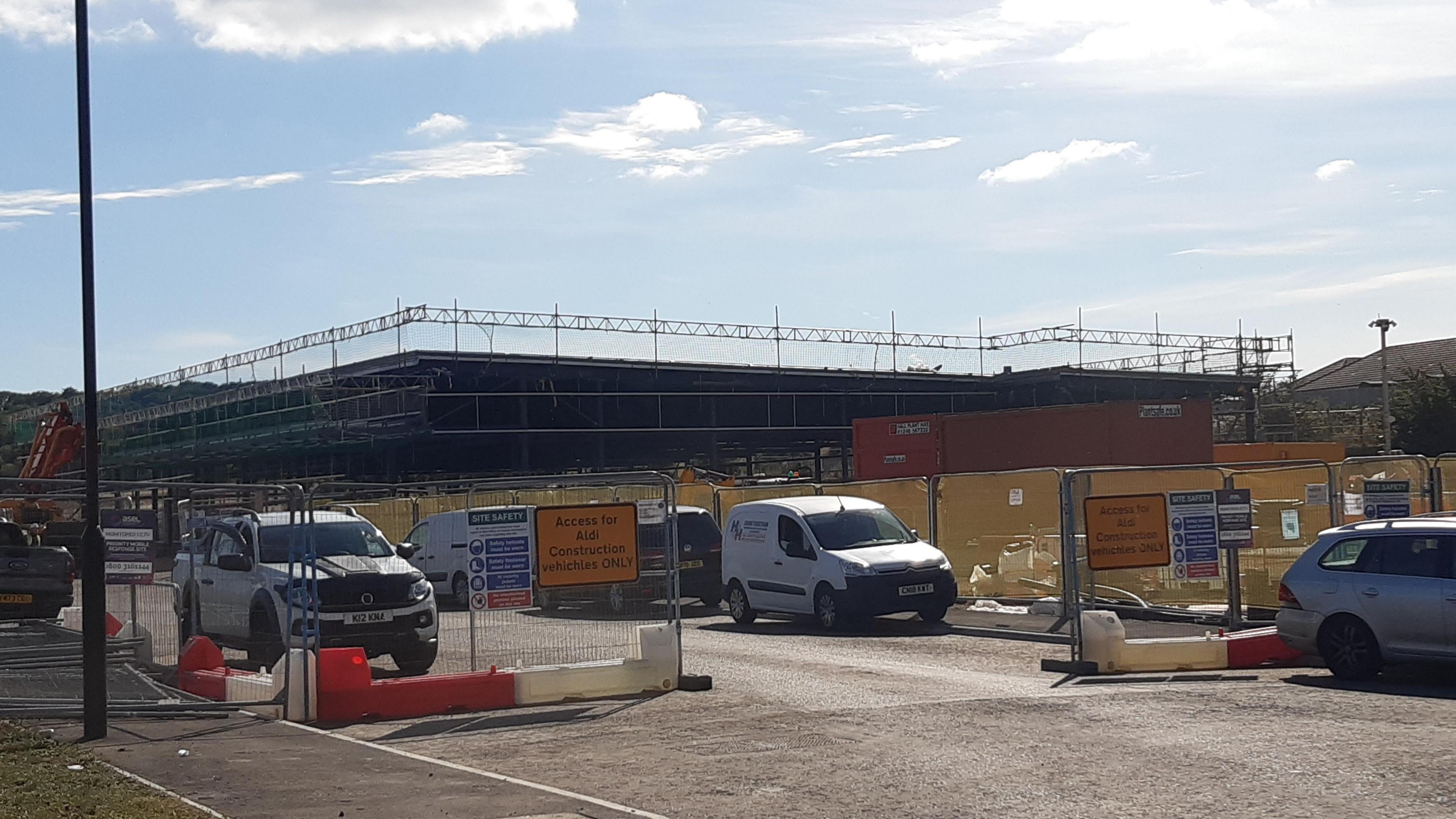 The new Aldi supermarket is under construction. The large warehouse is surrounded by scaffolding and fencing.