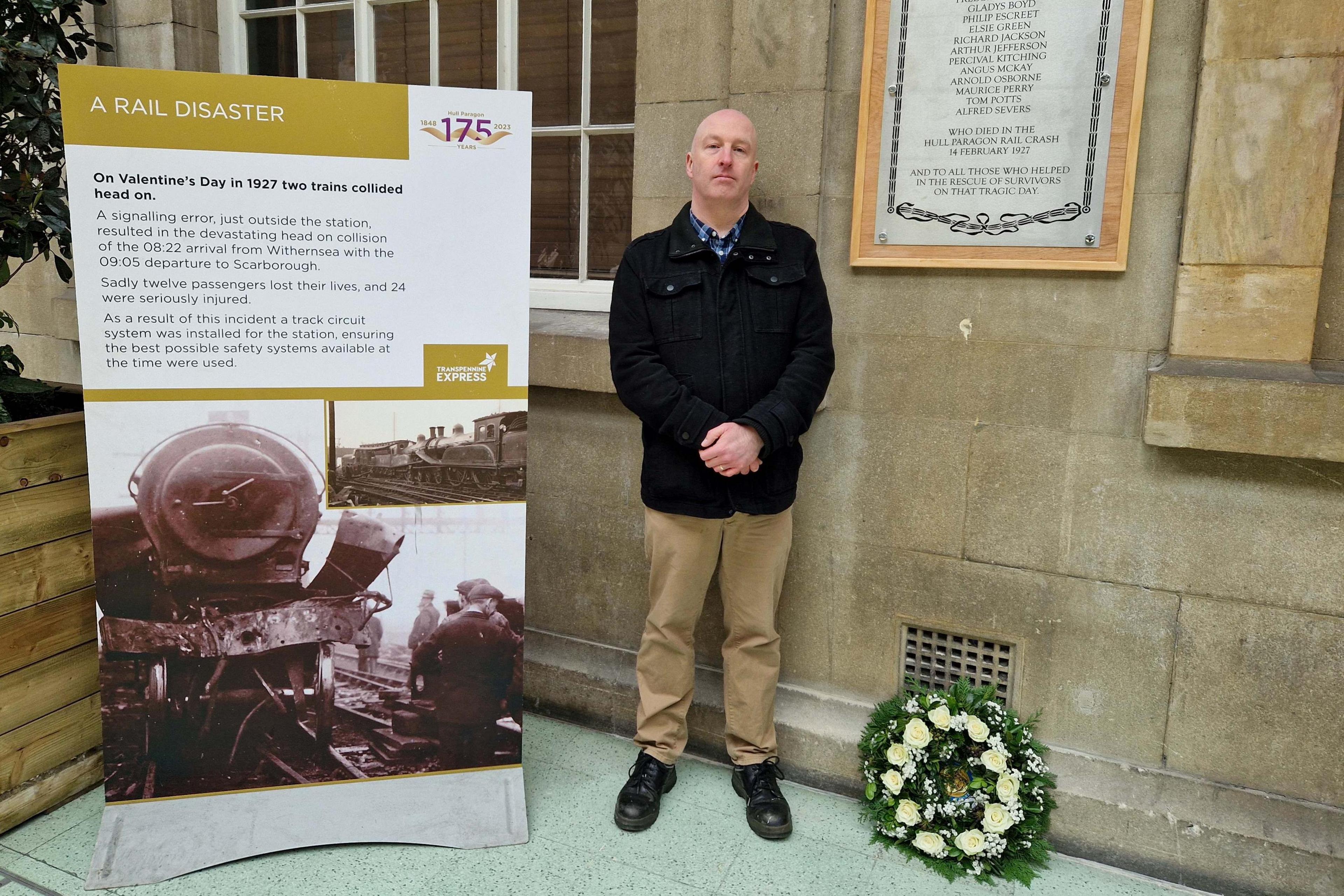 A man stands in front of a sandstone wall while wearing a chequered shirt, black jacket, beige trousers and black shoes. Next to him is a pull-up banner, of his height, with details of the Hull Rail Disaster, in black text on a white background, along with a black and white photo of a train engine. A floral wreath is on the floor to the right.