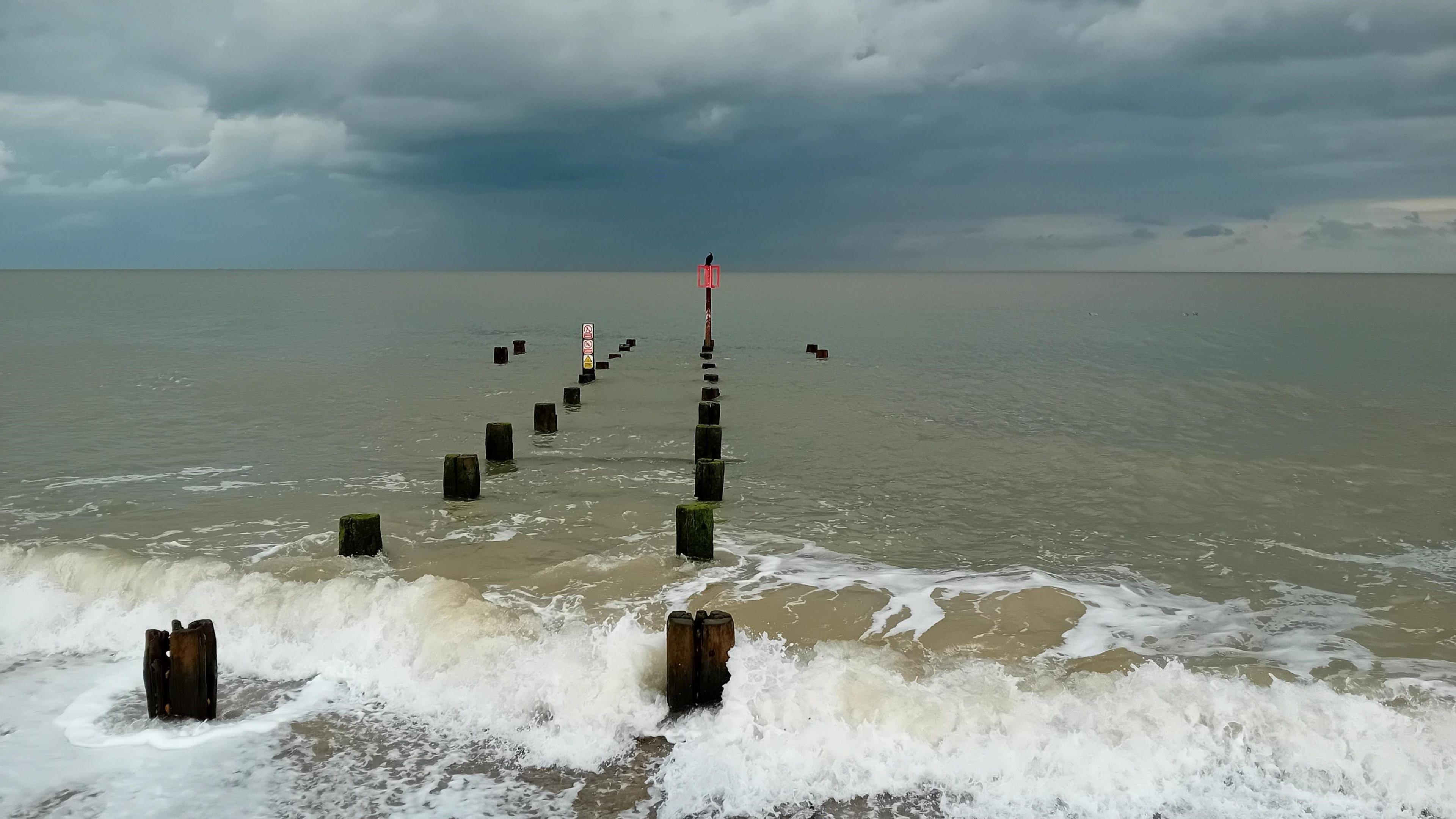 Two rows of wooden groynes stretch out to sea under a cloudy grey sky