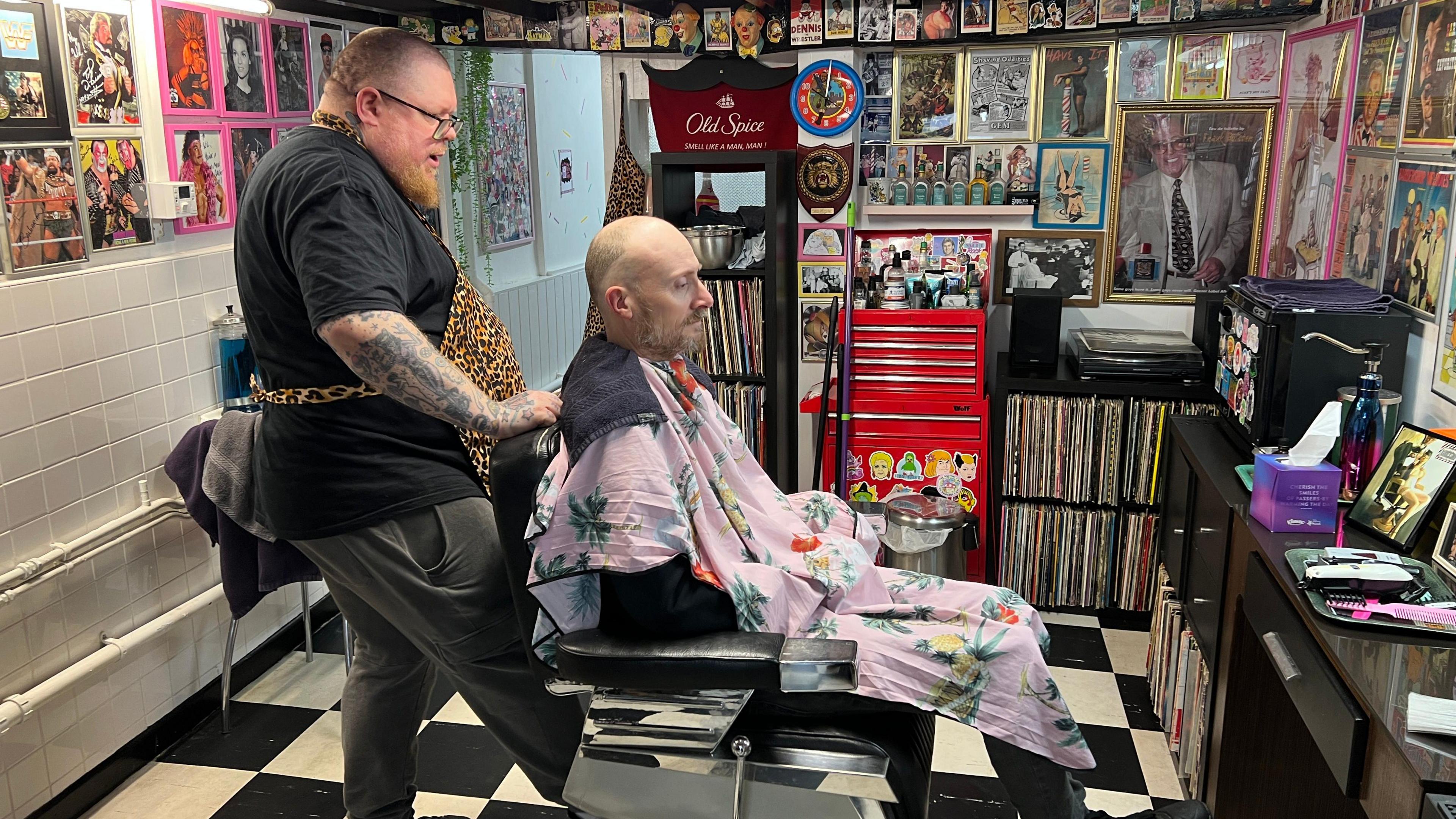 A man sits in a barbers chair with a pink gown. His chair is being adjusted by the barber standing behind him.