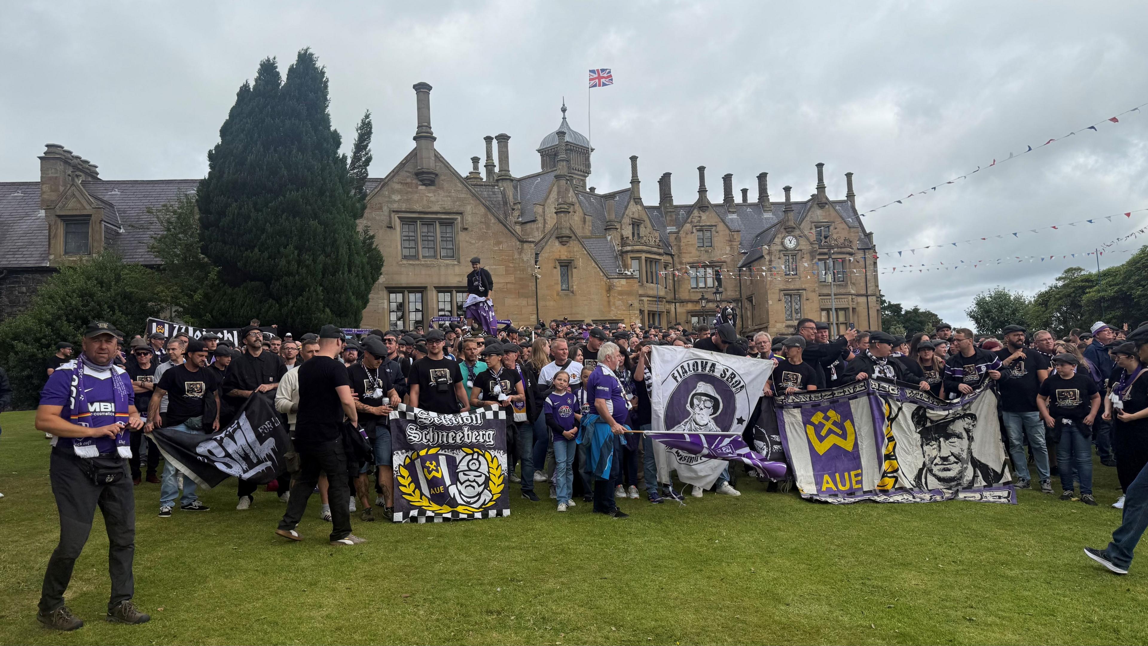 A large group of Erzgebirge Aue fans holding flags and banners standing outside Brownlow House - a brown brick building in a Baronial style