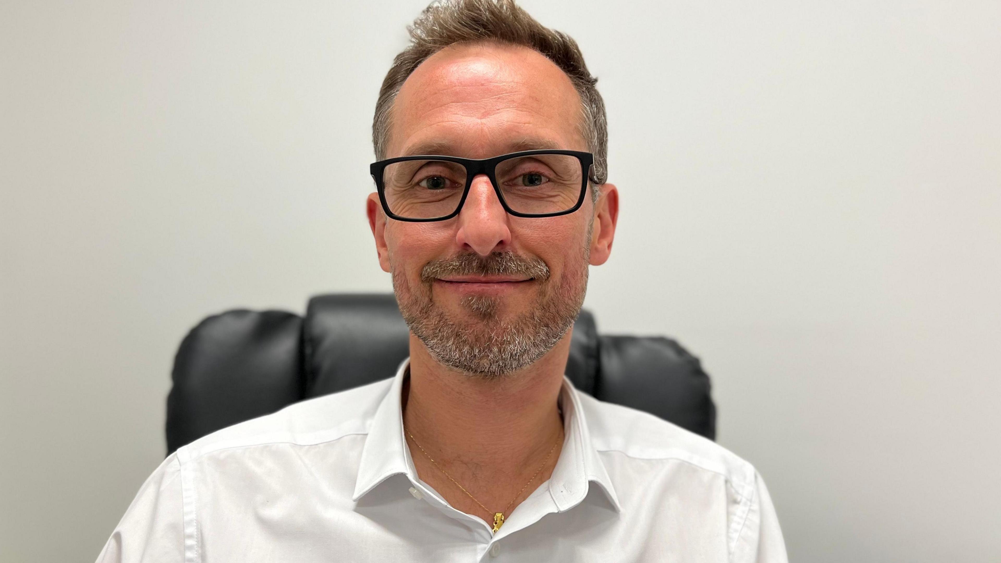 Flo Aranda is wearing a white shirt and glasses. He is sat on a black chair in an office. There is a white wall behind him. 