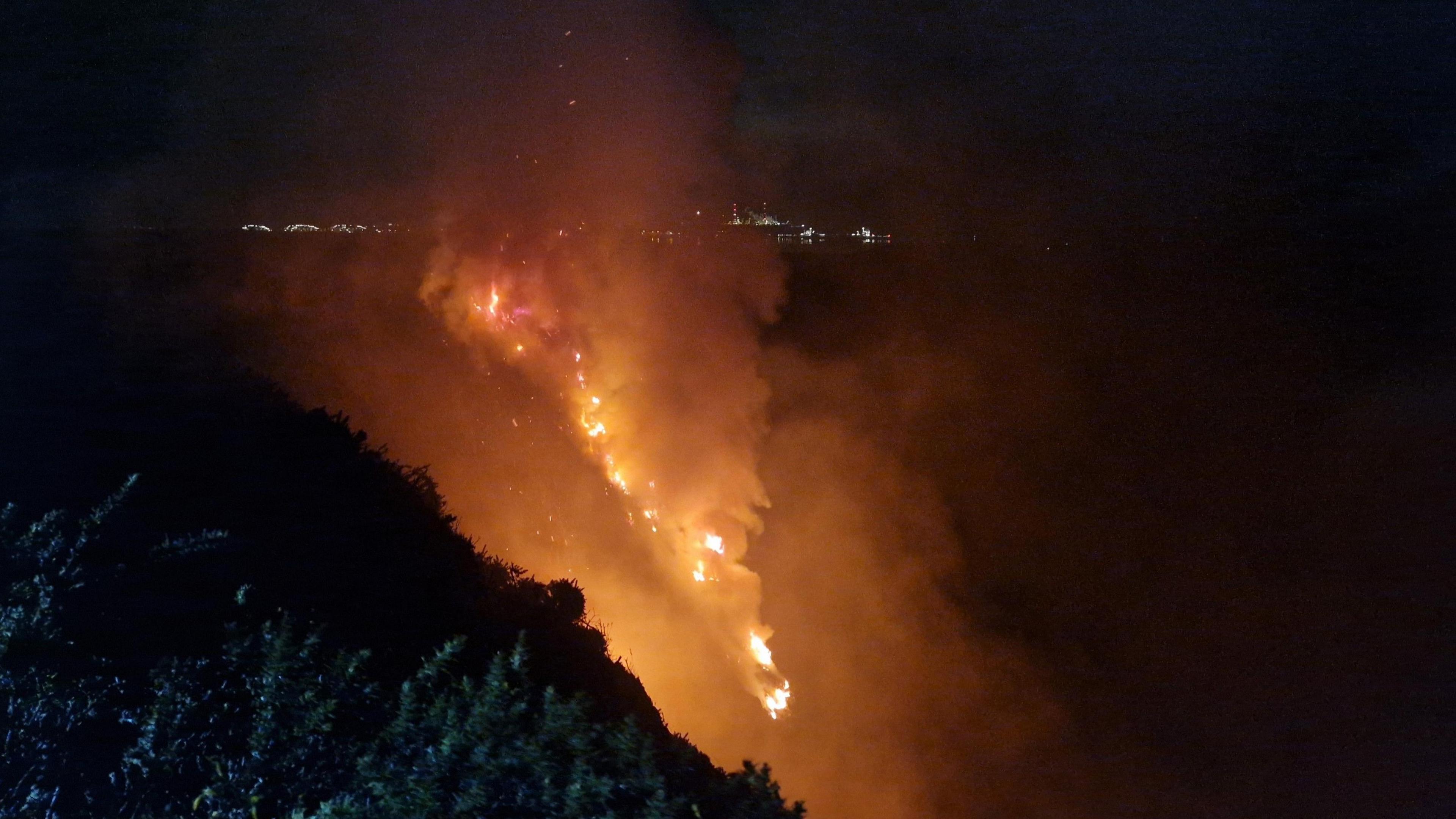 A large fire is scene at night on a hillside, with the lights of a town in the distance behind. 