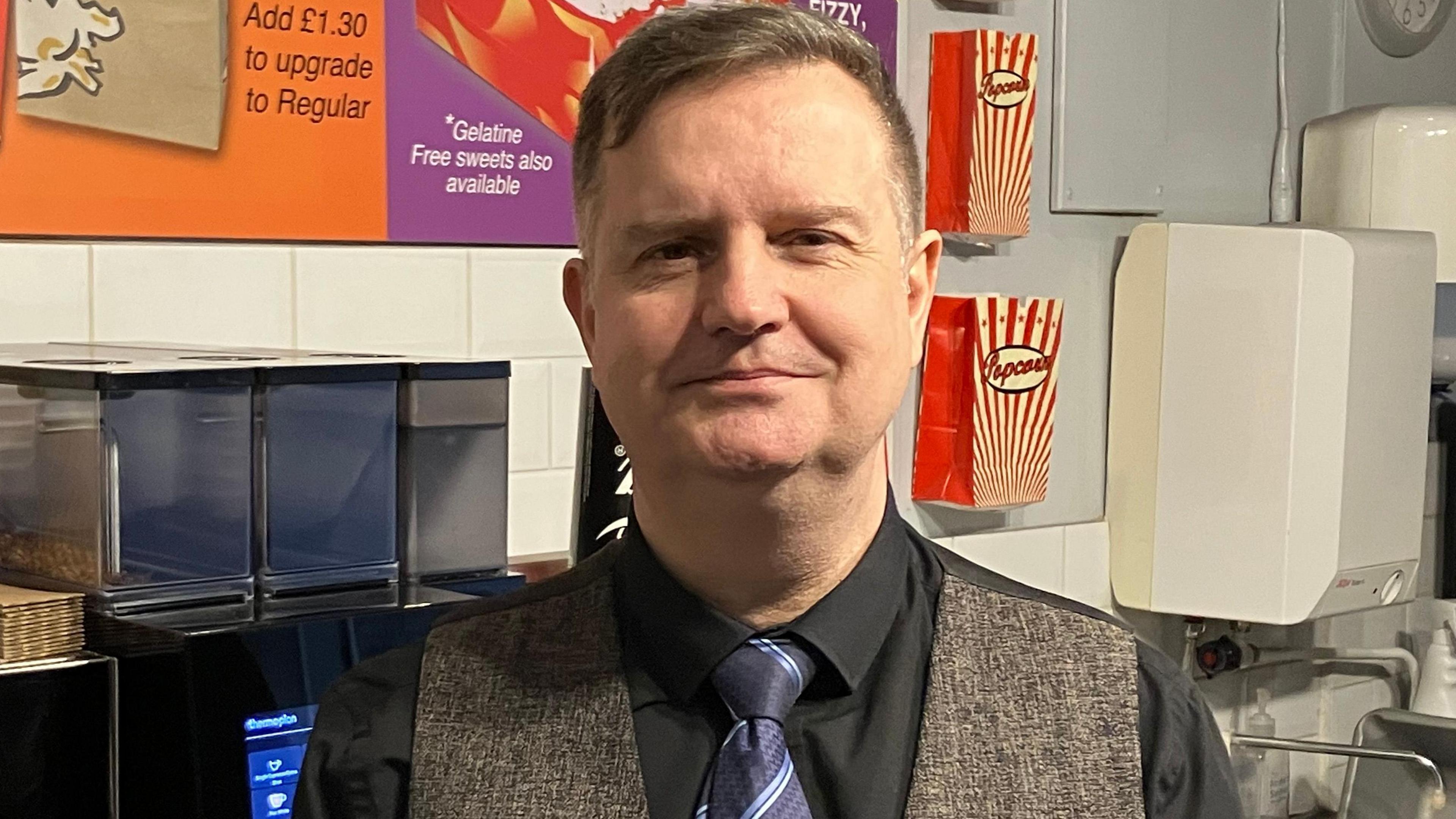 Michael Hansell stands in front of the snack bar area at the cinema wearing a grey waistcoat, black shirt and blue tie.