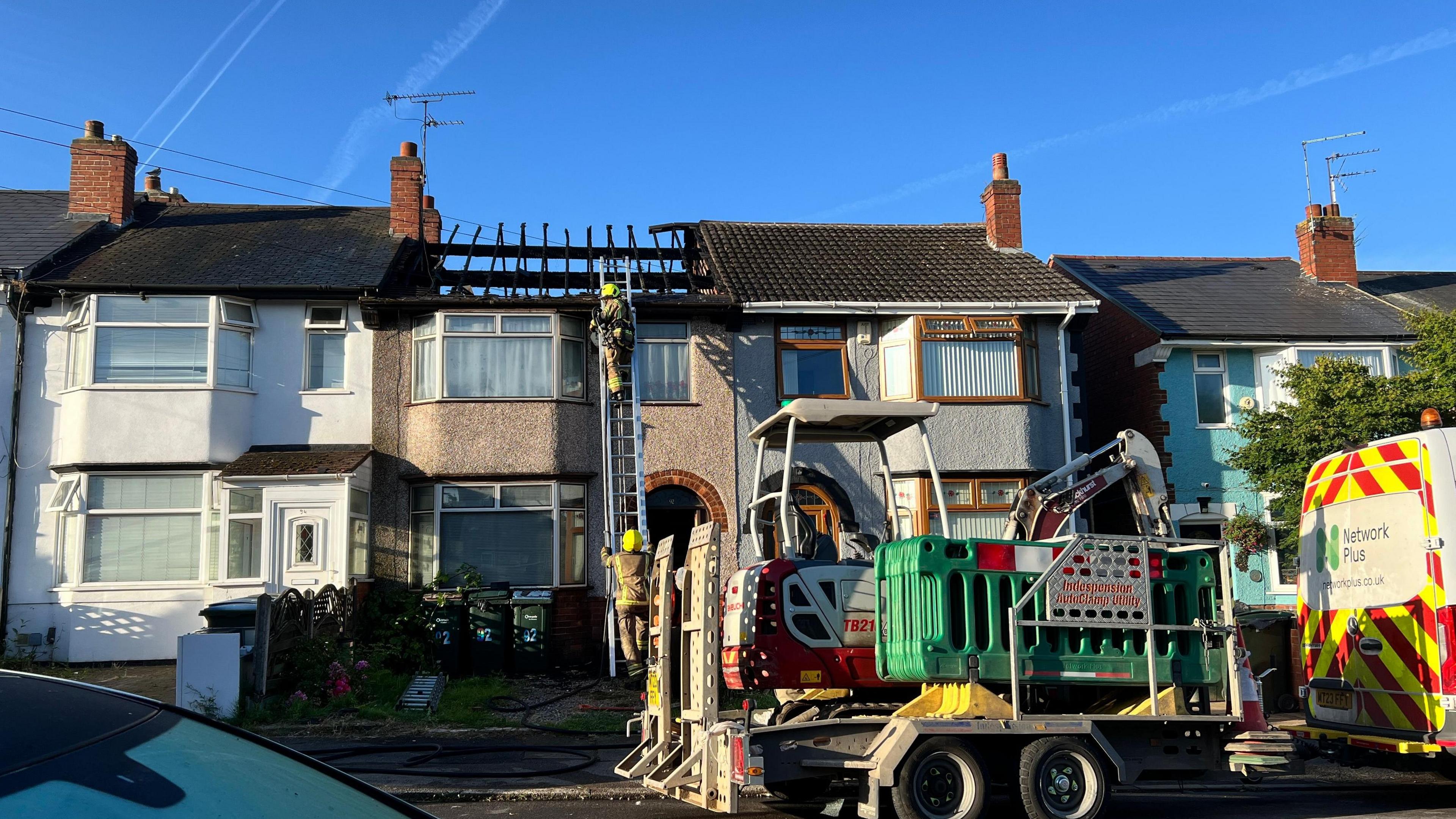 Terraced homes on a street. One of the homes has a burnt out roof and firefighters are climbing the house on a ladder