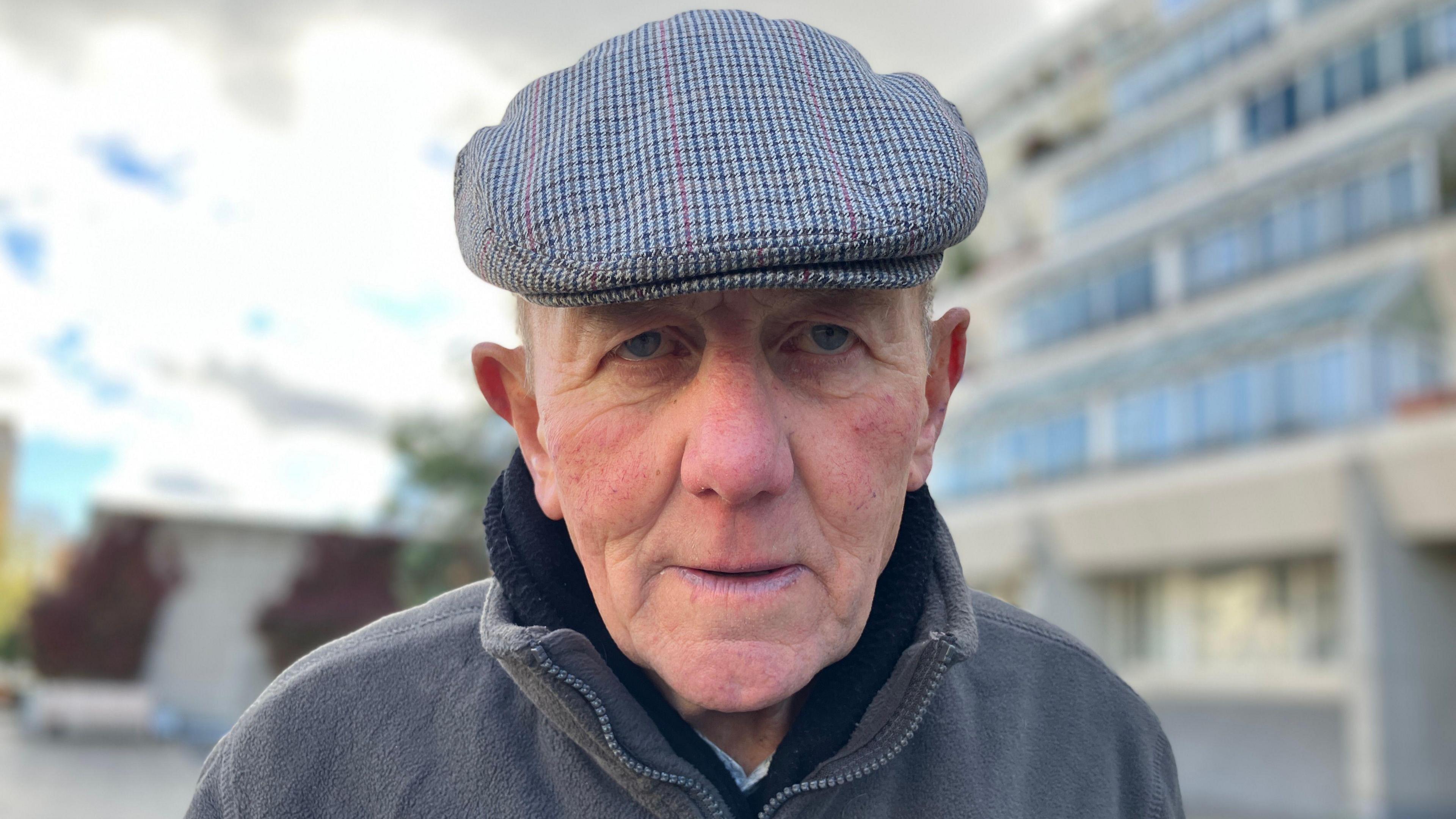 John Mahoney in a fleece jacket and a blue and red patterned flat cap.