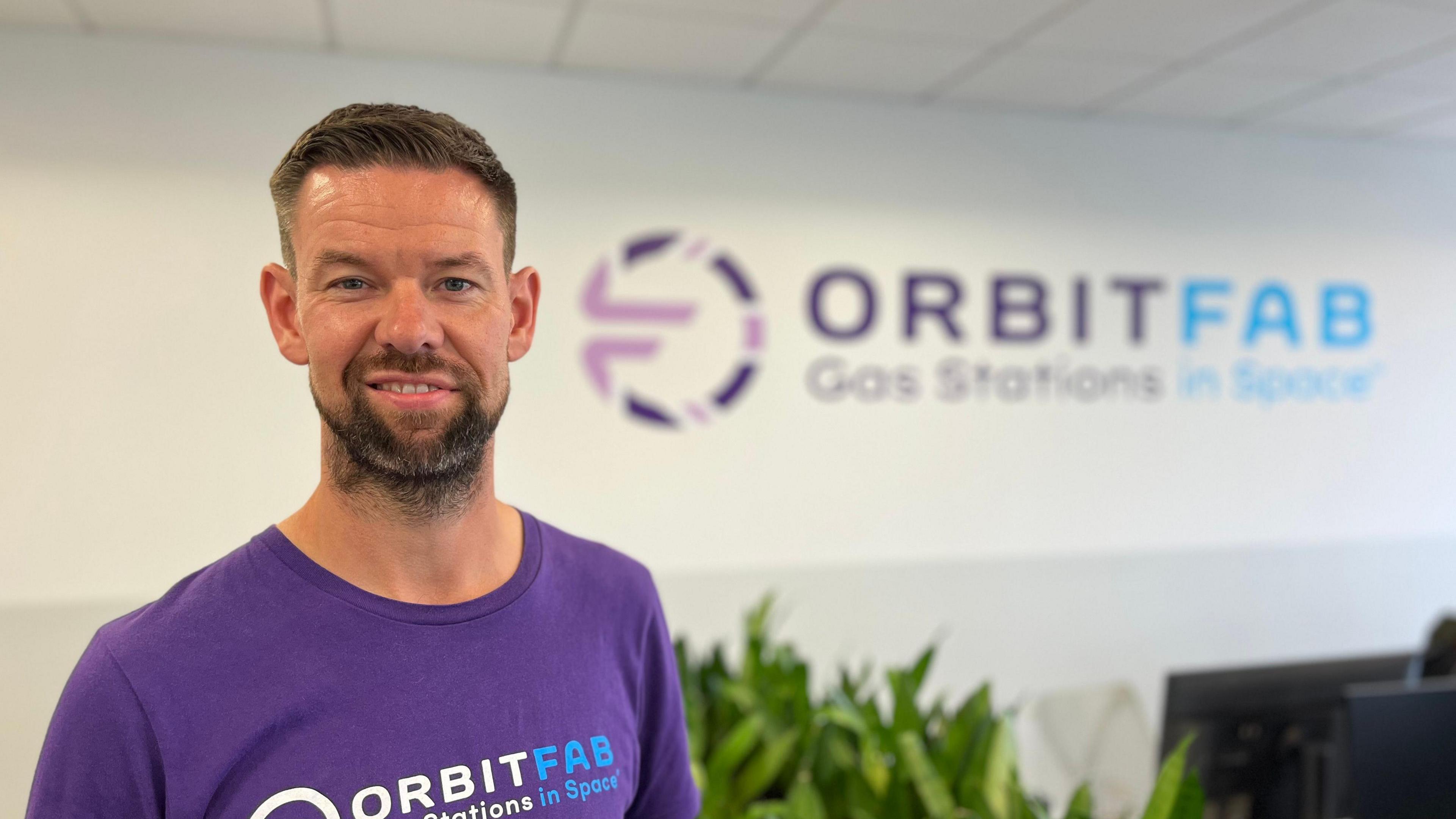 Jacob Geer, wearing a purple t-shirt with the words 'Orbit fab' on in blue and white writing. He has short brown hair and brown stubble. Behind him is a white wall with the same logo on as his t-shirt.