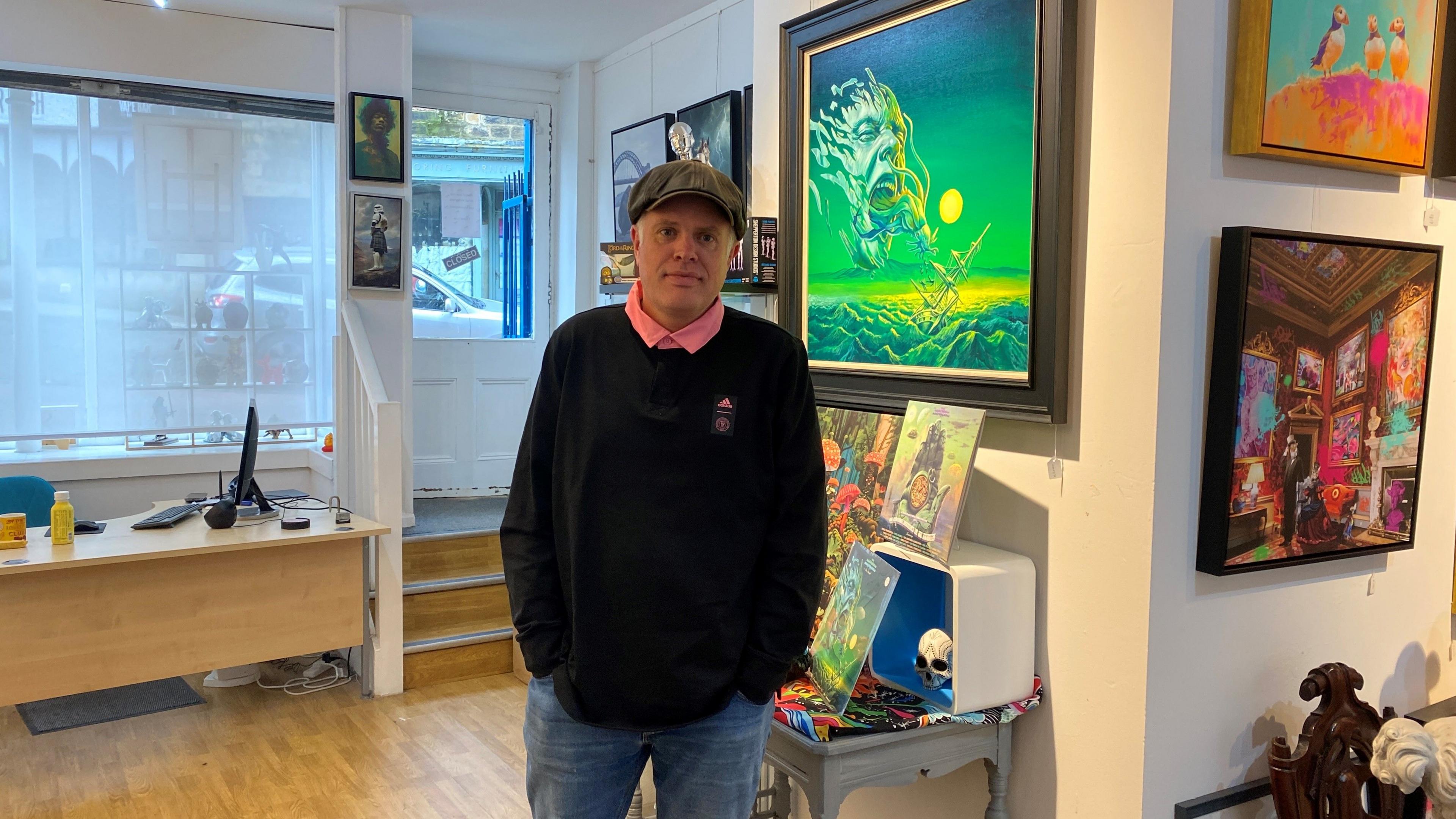 A man wearing a cloth cap stands in front of pictures of art in a gallery.