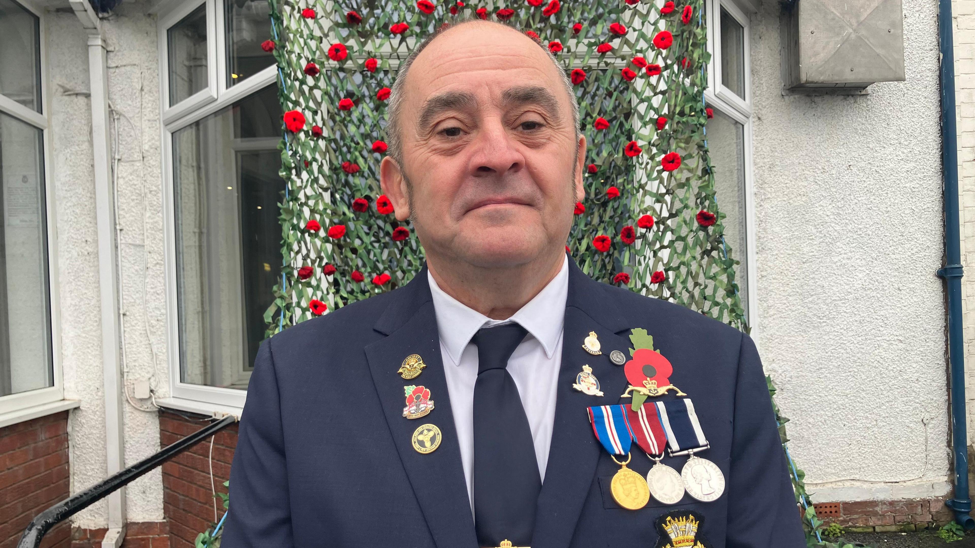 Veteran Darren Hardy dressed in a blue jacket displaying a poppy and his medals. He is standing in front of a poppy display and looking directly towards the camera.