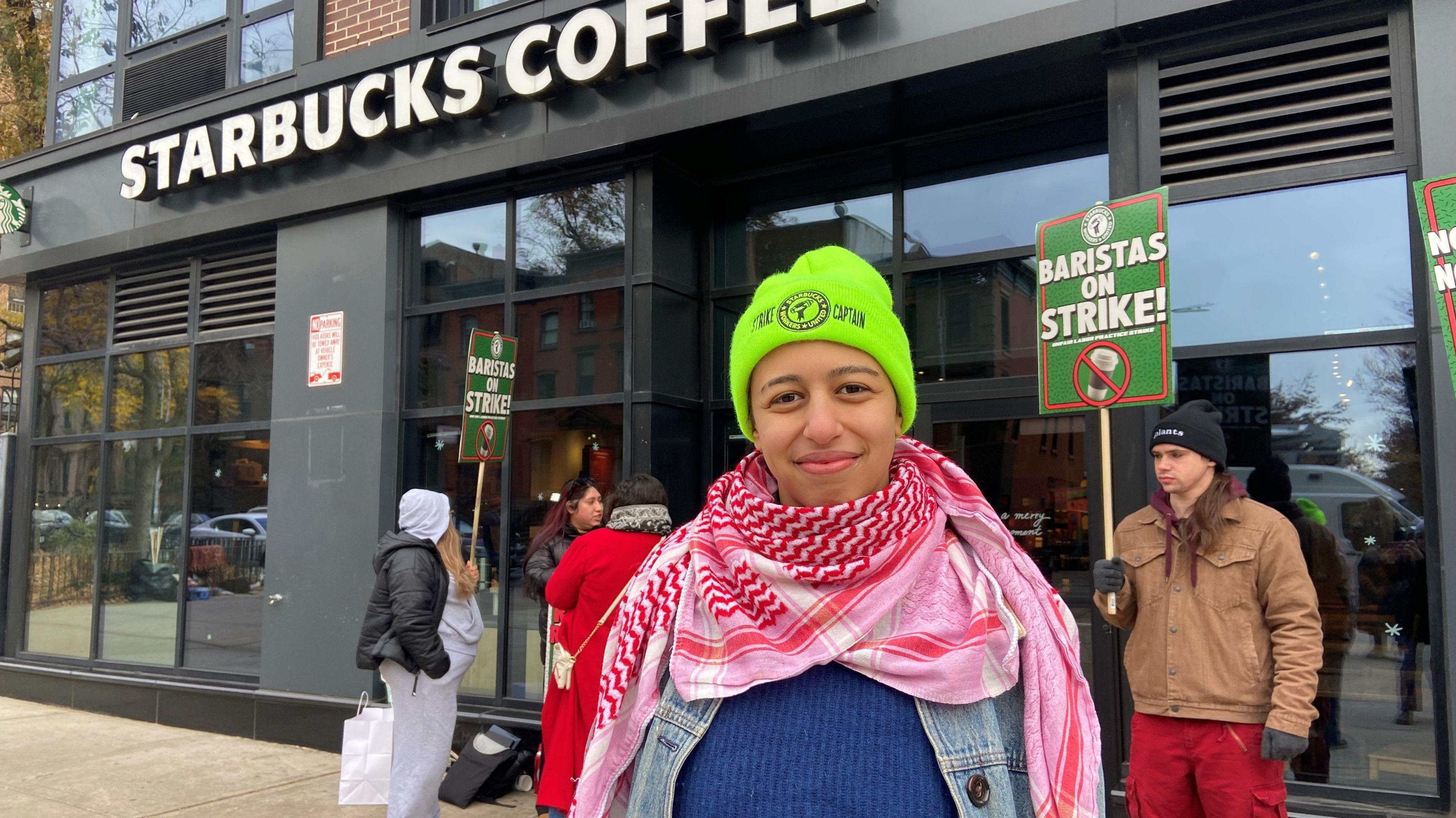 A woman wearing a neon beanie stands in front of a Starbucks coffee shop in Brooklyn, New York, alongside people holding signs reading "Baristas on Strike".