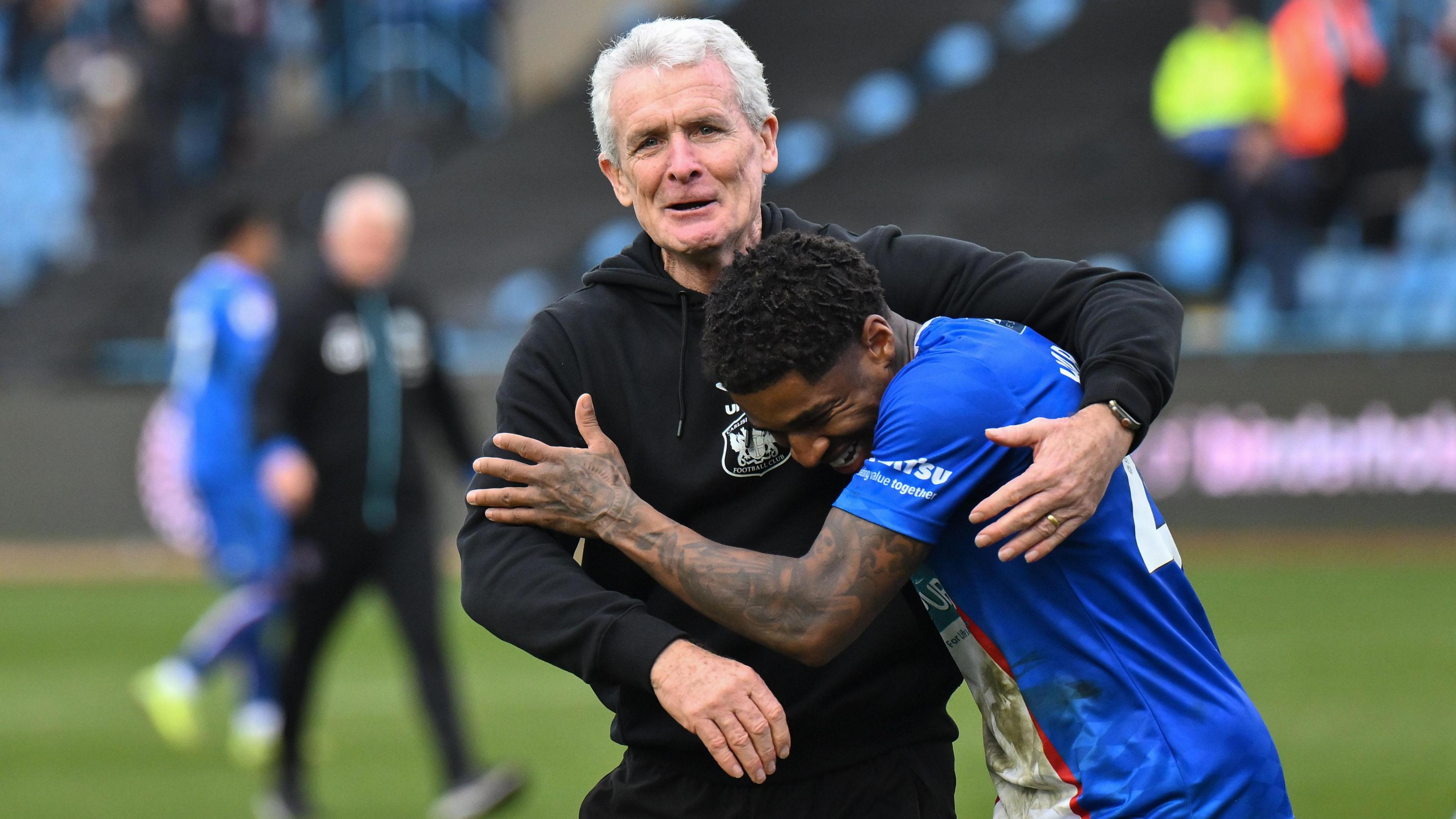 Carlisle manager Mark Hughes in his black tracksuit hugs his player Kadeem Harris at full-time after a crucial win