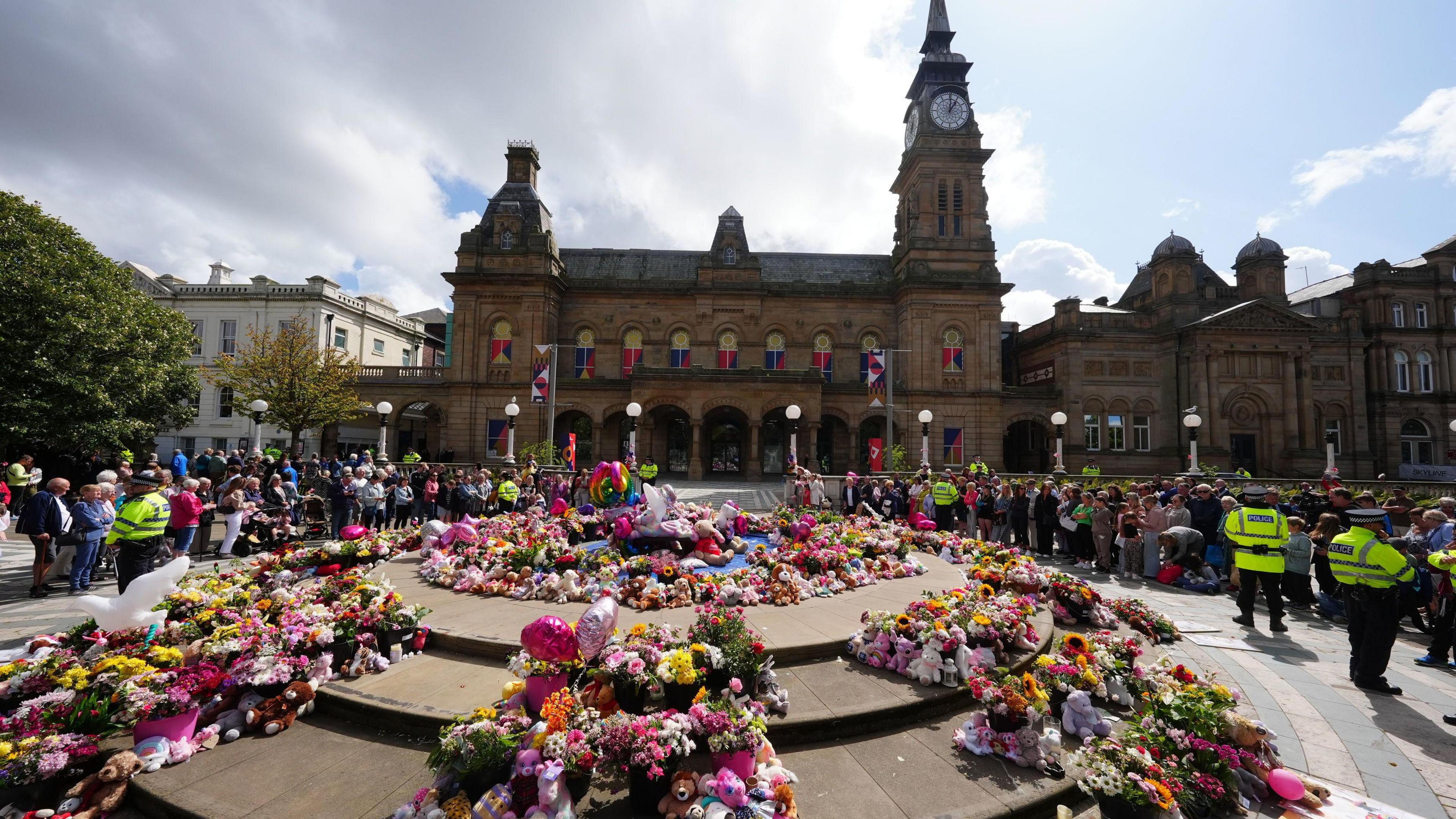 Floral tributes in a square outside The Atkinson arts centre in central Southport,  with large displays left on a circular stone area in the middle of a square in front of an ornate Victorian building in stone, with a clocktower at one end. Peaceful crowds are being marshalled by a handful of police officers 