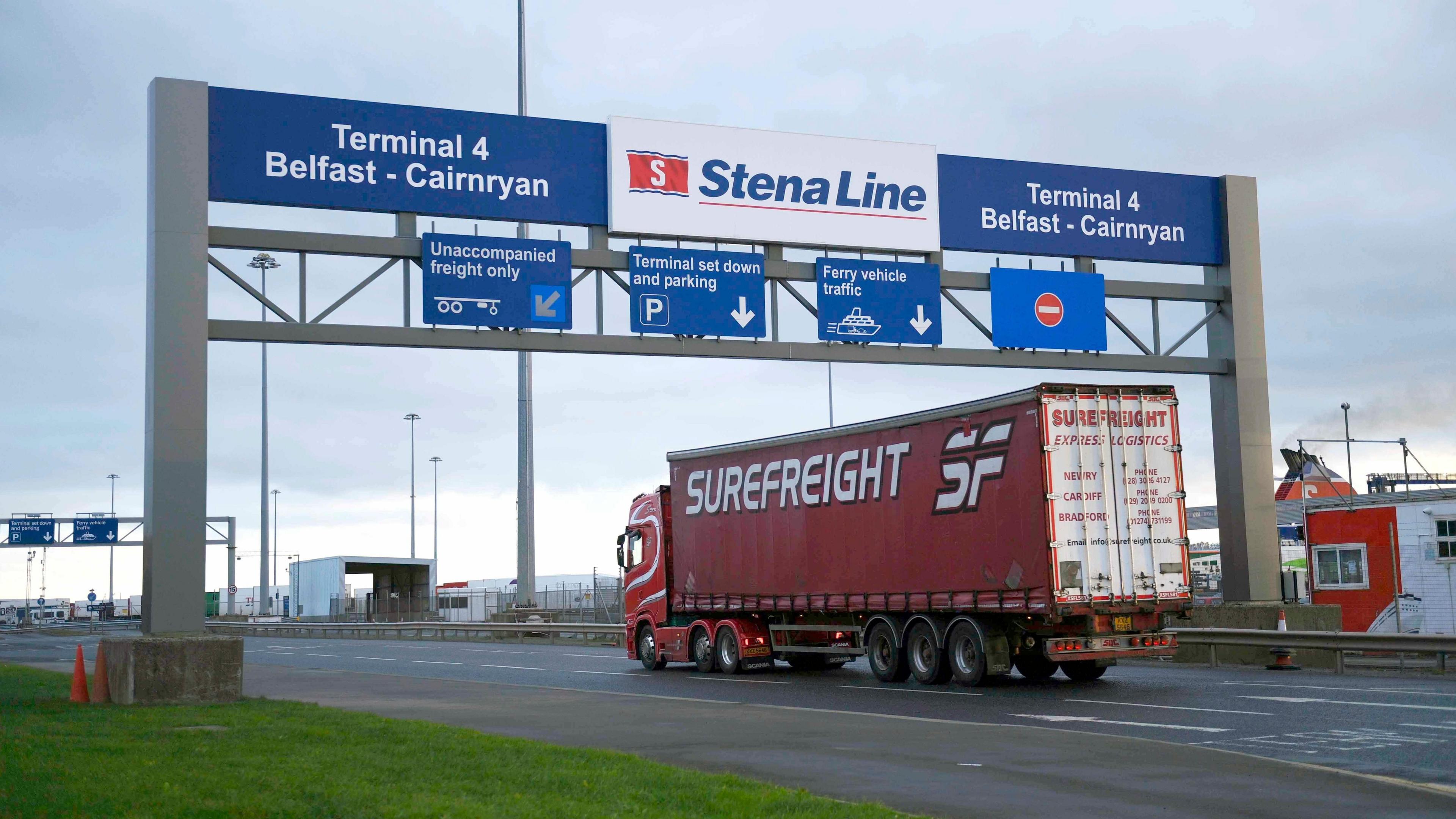 A freight lorry travelling through the Port of Belfast. The lorry is red with 'superfreight' on it side in white letters. The back doors of the lorry are white. It is driving under a white Stena Line sign, the sign also says terminal 4, Belfast- Cairnryan. The sign is blue with white writing. 