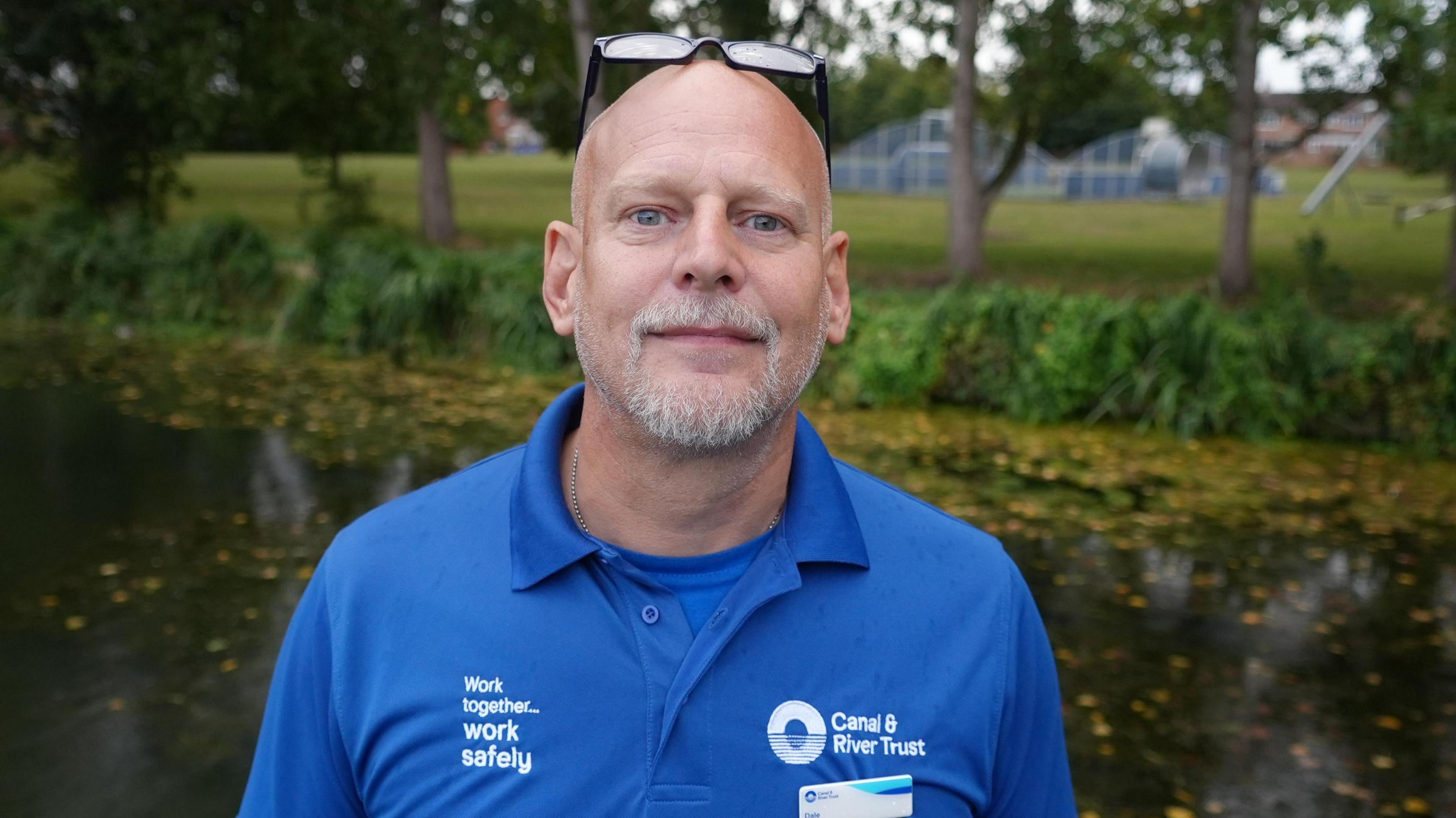 A man standing by a river wearing a blue shirt with a Canal & River Trust logo, and sunglasses on his head.
