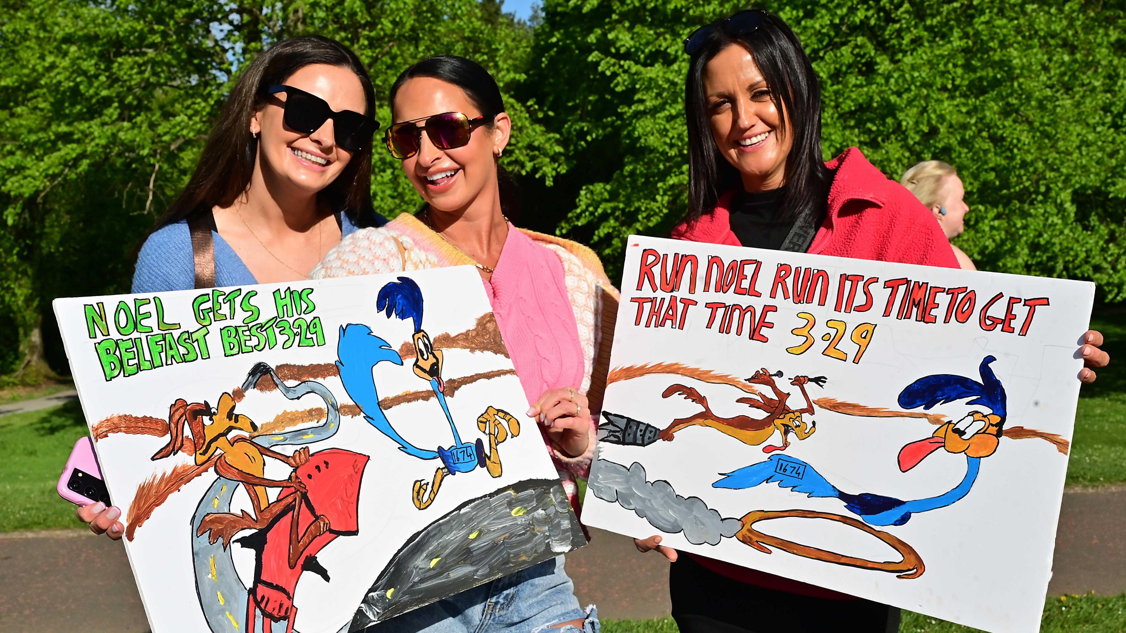 Three ladies are holding up signs to support those who are running. They all have dark hair and are wearing sunglasses with blue, pink and red clothing.