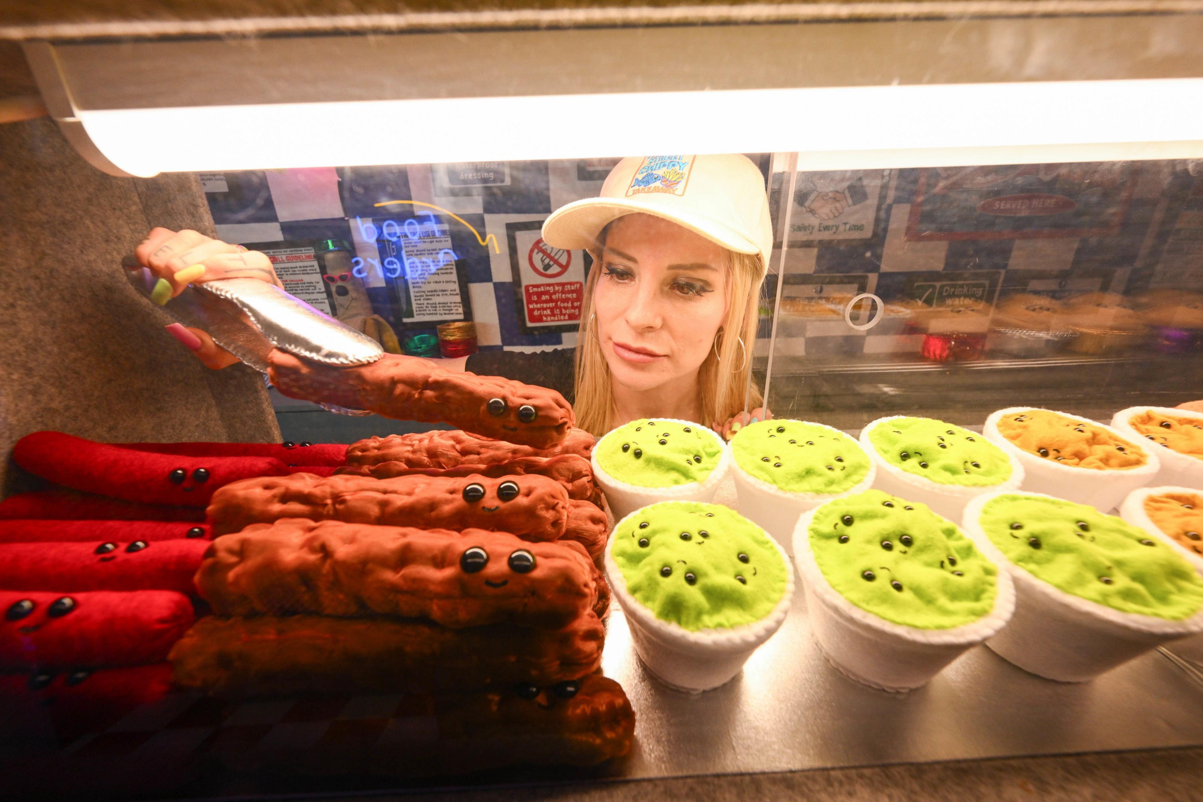 Artist Lucy Sparrow picks up a felt sausage from behind the chip-shop counter using tongs. There are also small felt pots of mushy peas behind the counter.