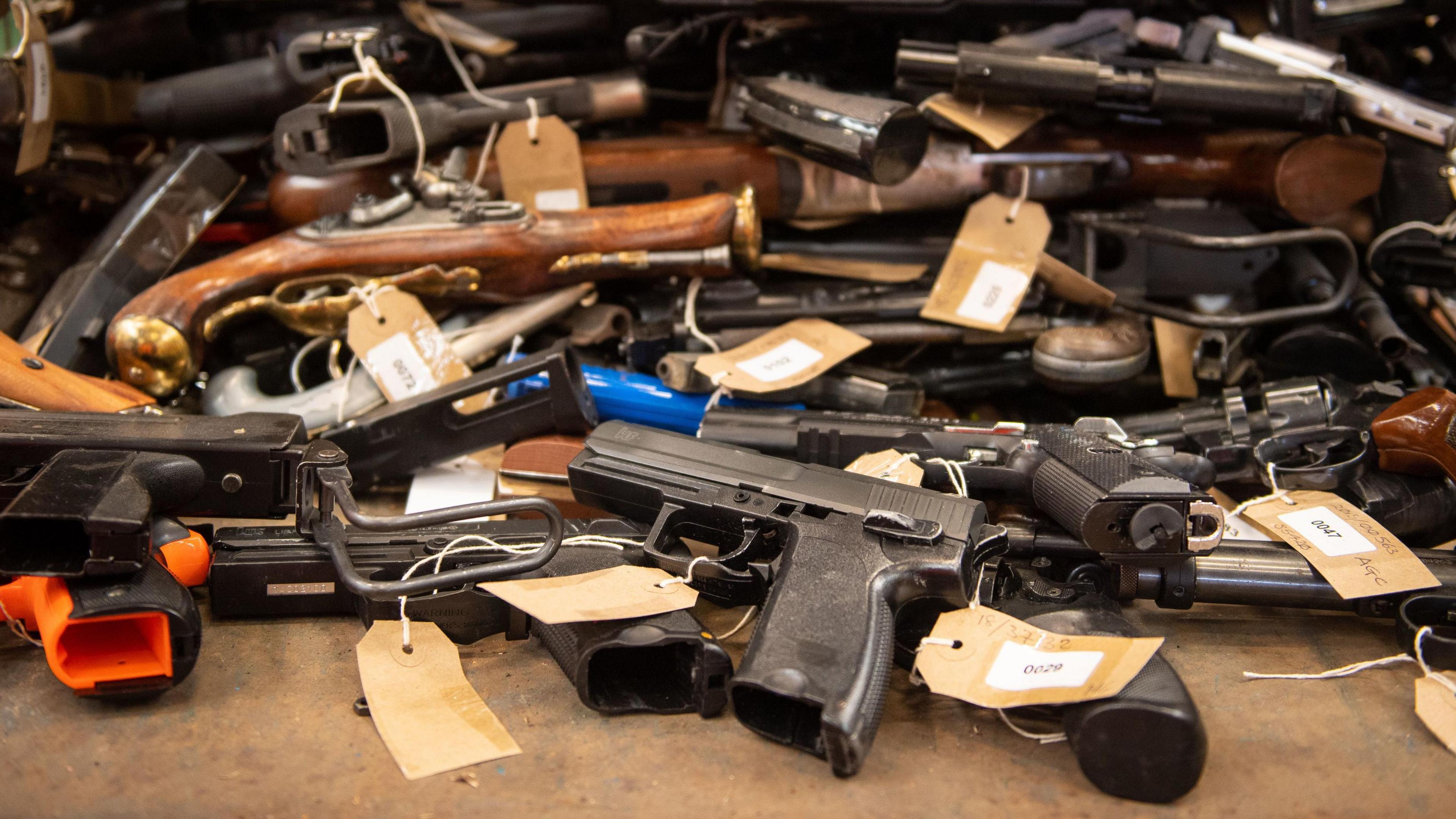 A large collection of guns labelled with brown paper luggage tags piled up in a room.