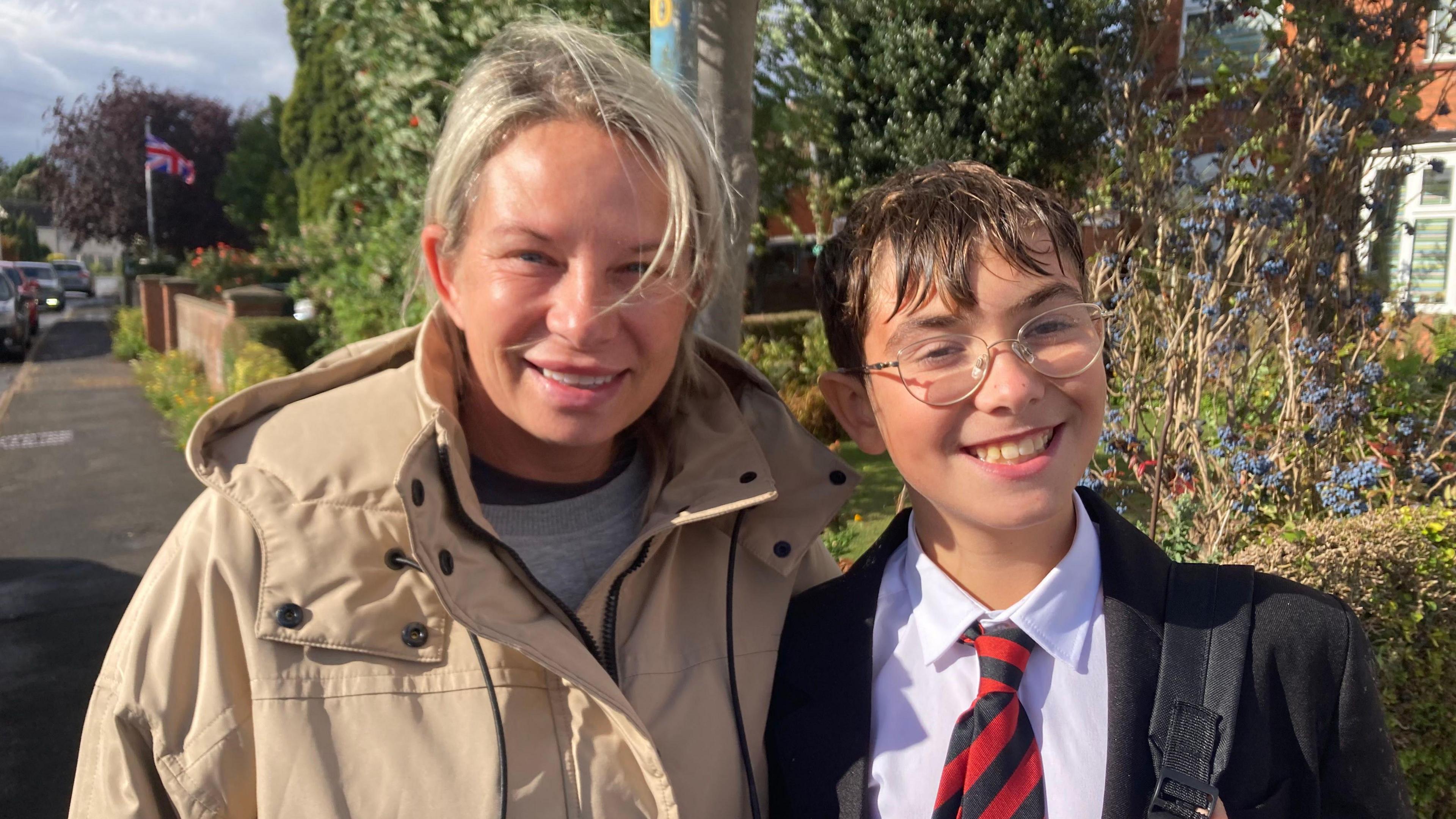 A head and shoulders shot of a woman - Izabela Santos - and a young boy -Leandro Santos - smiling at the camera. The woman has blonde hair tied back and is wearing a cream-coloured rain coat. The boy is wearing a school uniform consisting of a white shirt, a black blazer and a red and black striped tie. He has short brown hair and is wearing glasses and a backpack. 