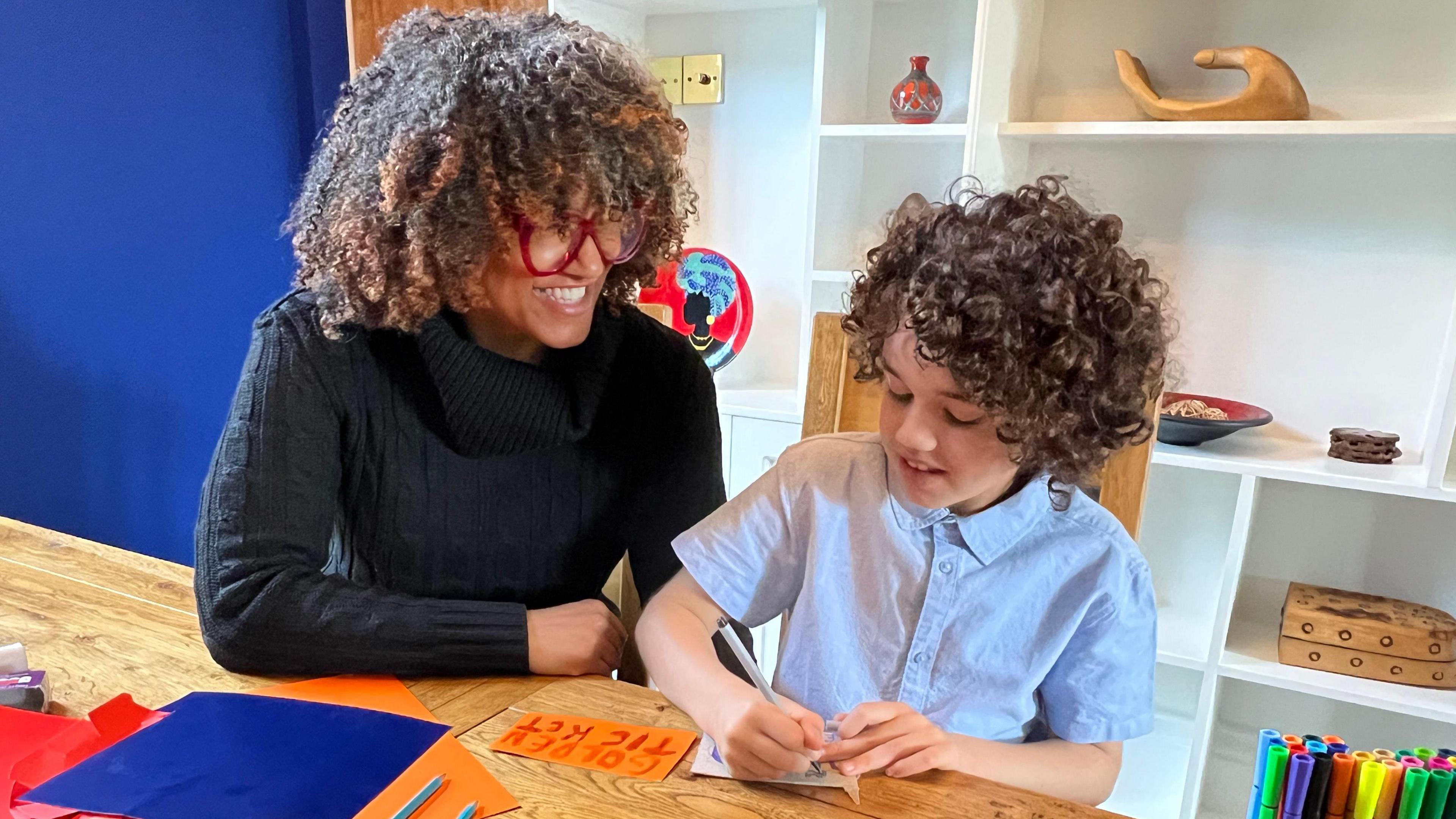 Christina Lipede at home with her son, aged nine. The pair are sitting at a table and her son is writing.