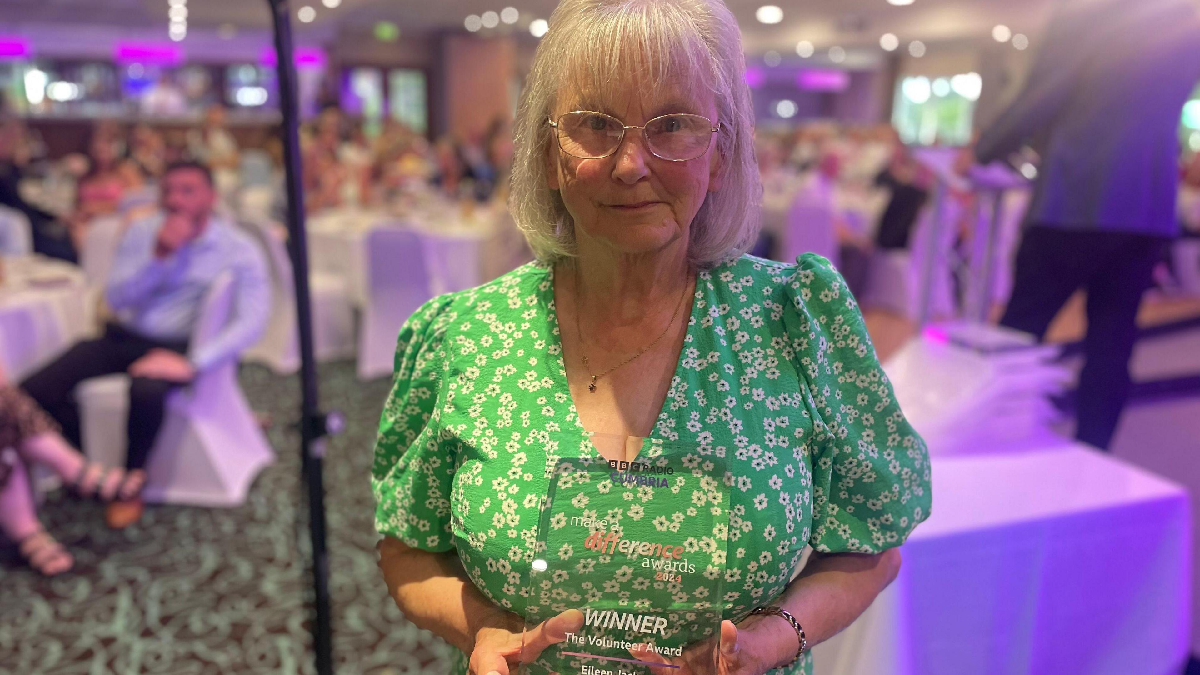 Eileen wearing a green dress in a ceremony hall holding her volunteer award.