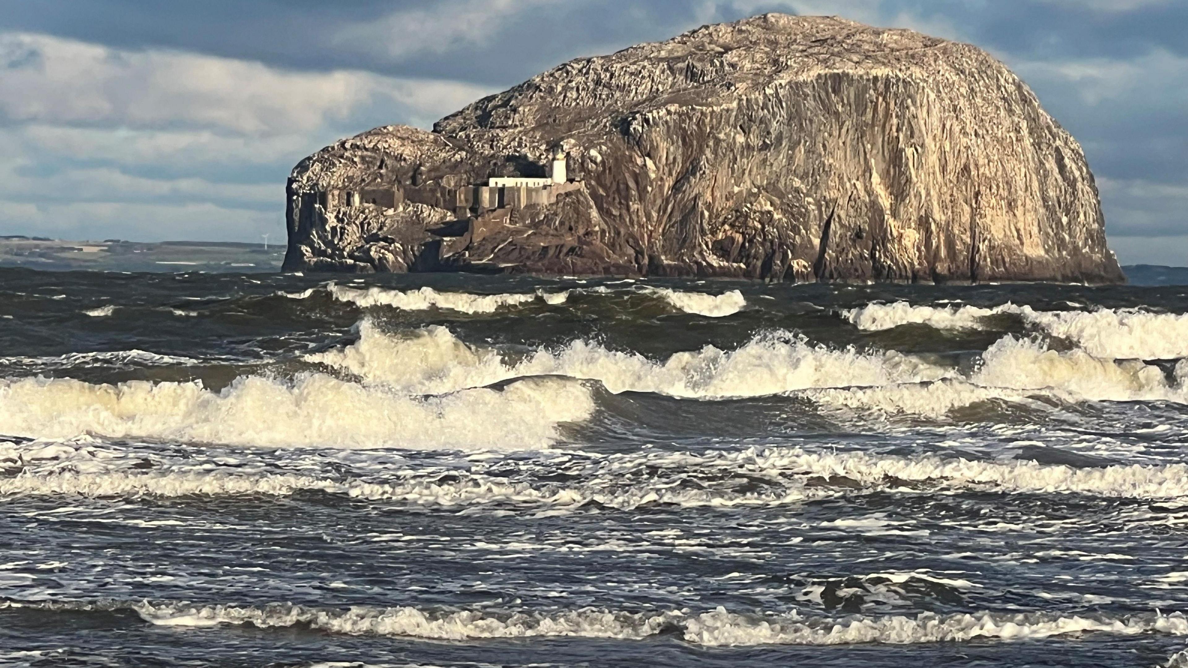 There is a lot of froth in the water and big waves. In the background is the Bass Rock.