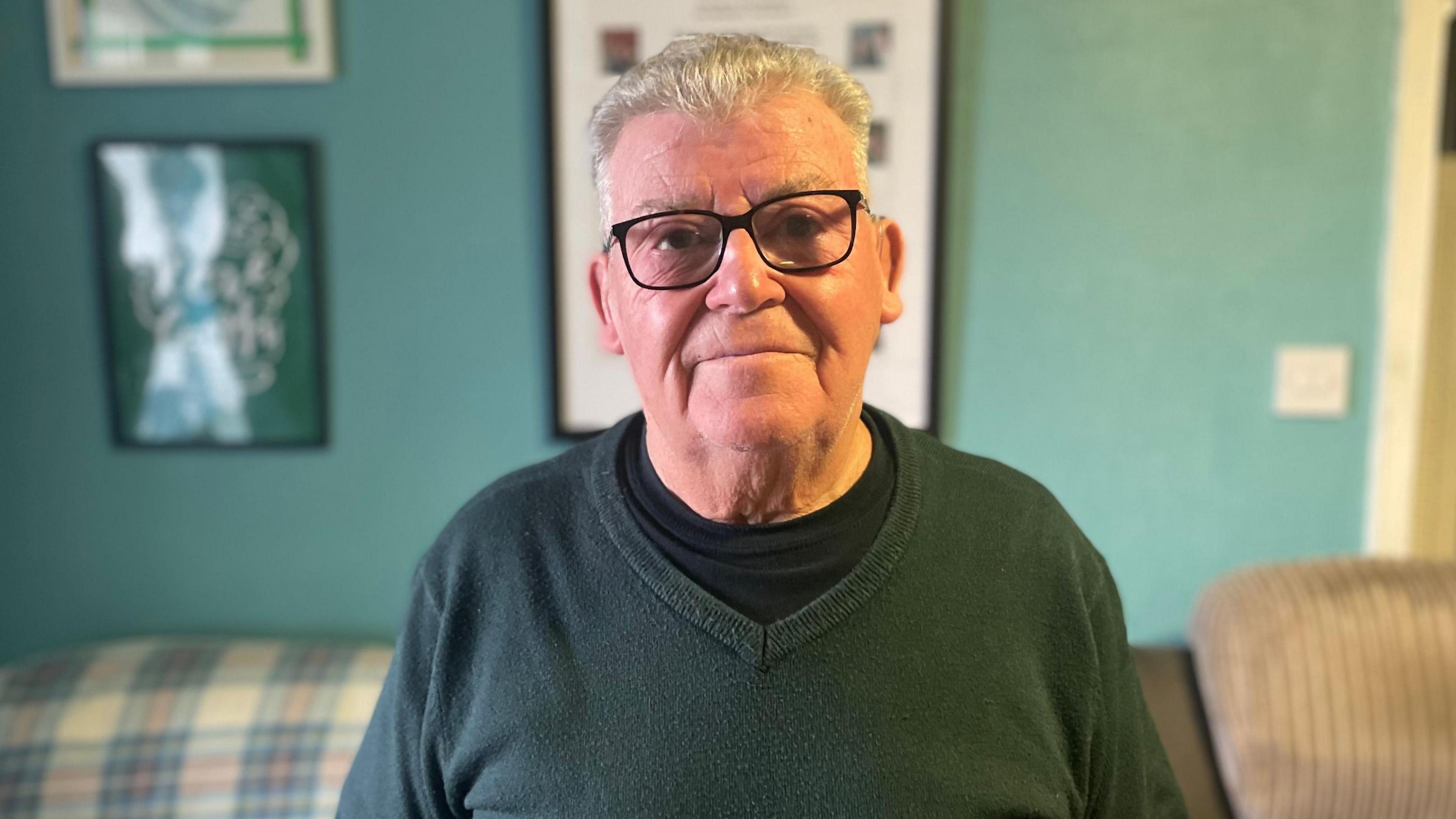 Allan Mackie looking straight at the camera. He is wearing a green jumper with a v-neck over a dark t-shirt with a crew neck. He is wearing dark-framed glasses. He has white hair. He is standing in front of a green background.