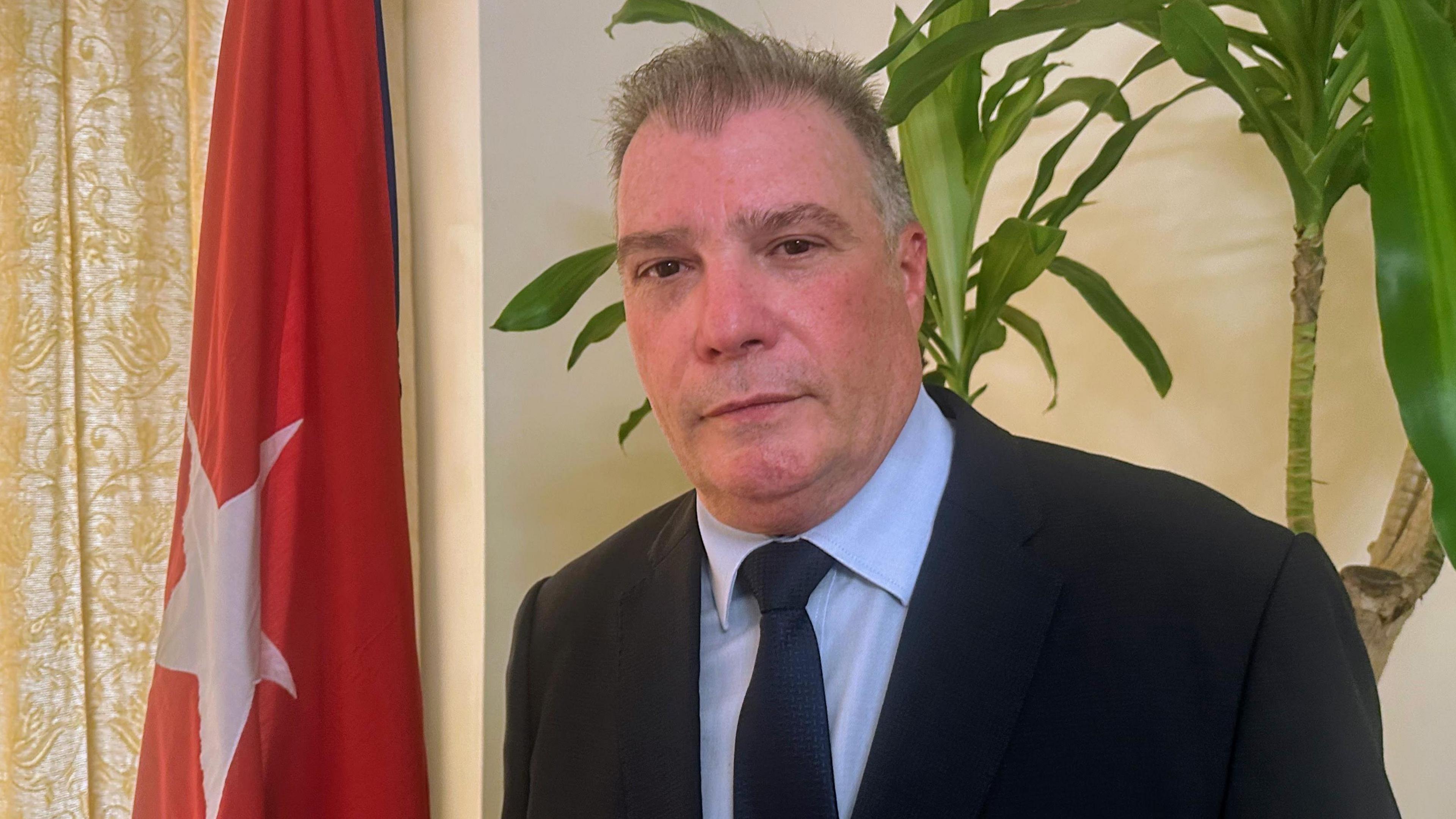 Juan Carlos García Granda stands in front of a Cuban flag. He is wearing a dark suit, a navy tie and a light blue shirt.