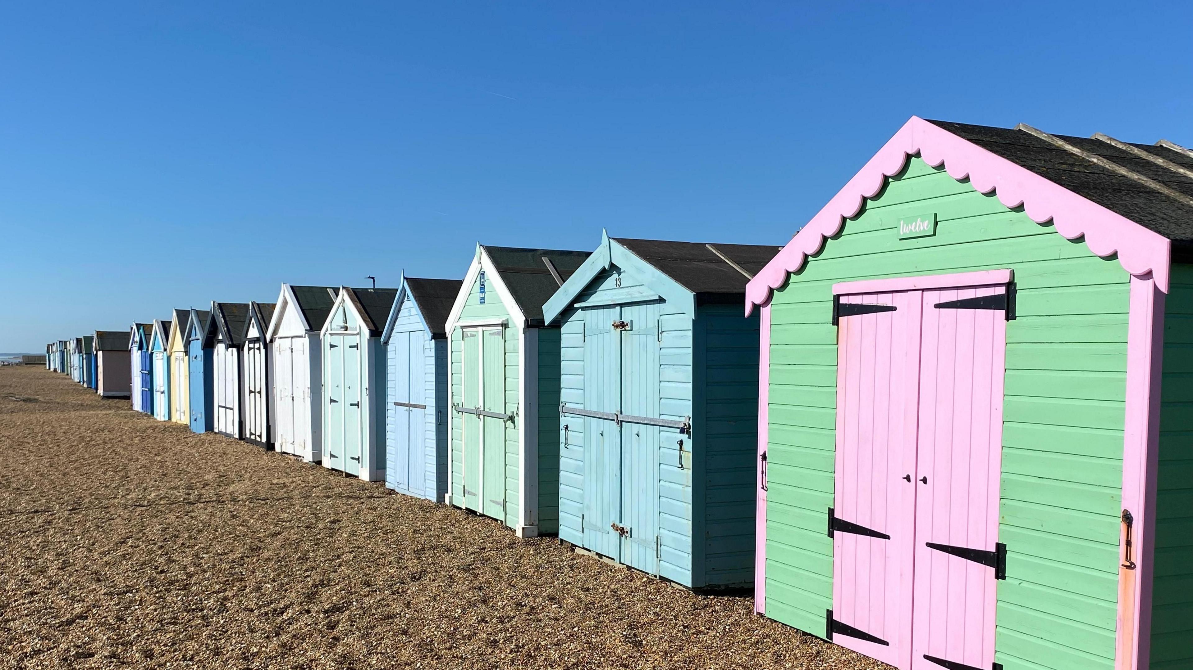 Coloured beach huts on a gravel beach, below a blue sky