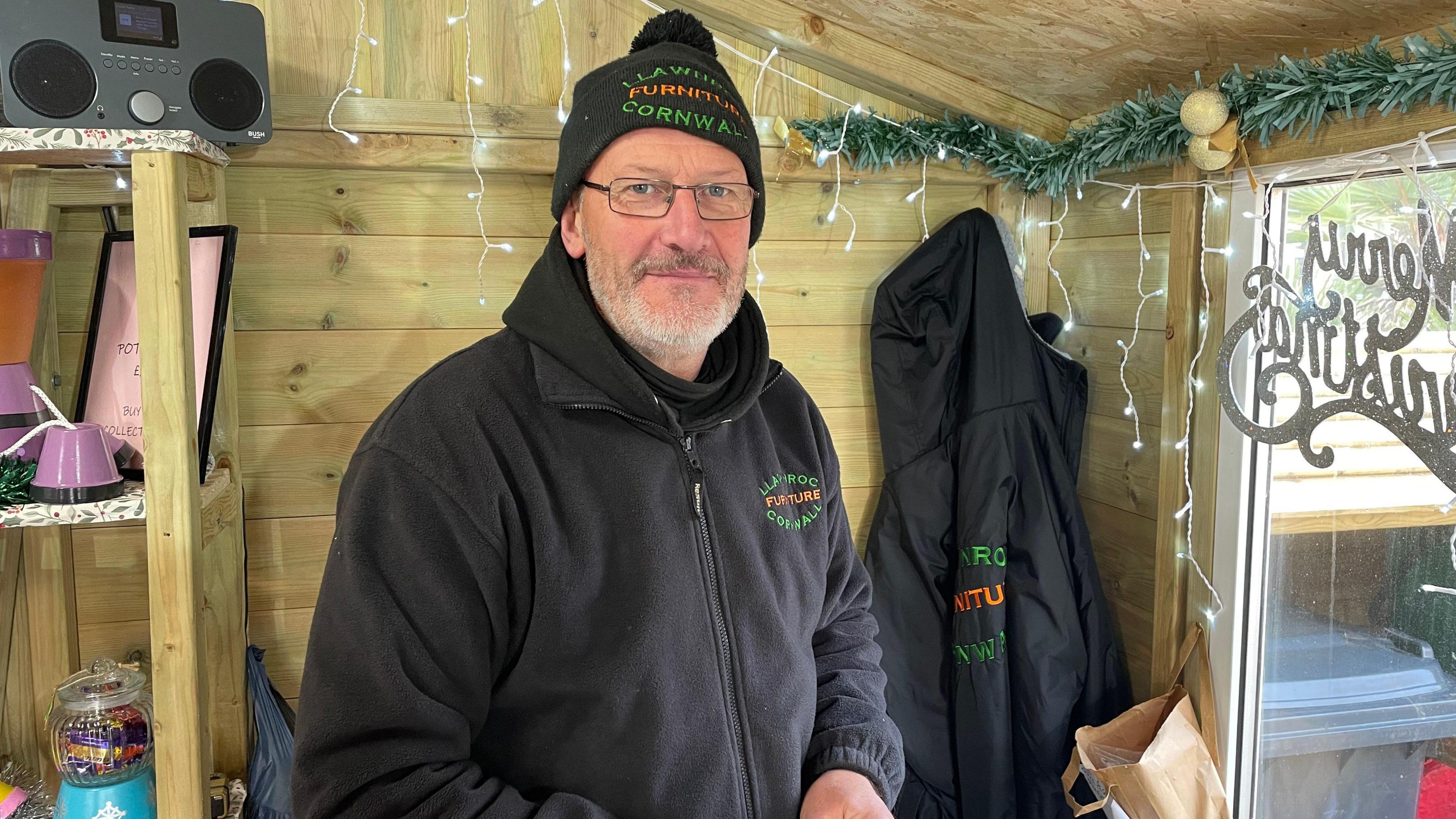 A man in a black jumper with black beanie in a wooden shed with Christmas decorations