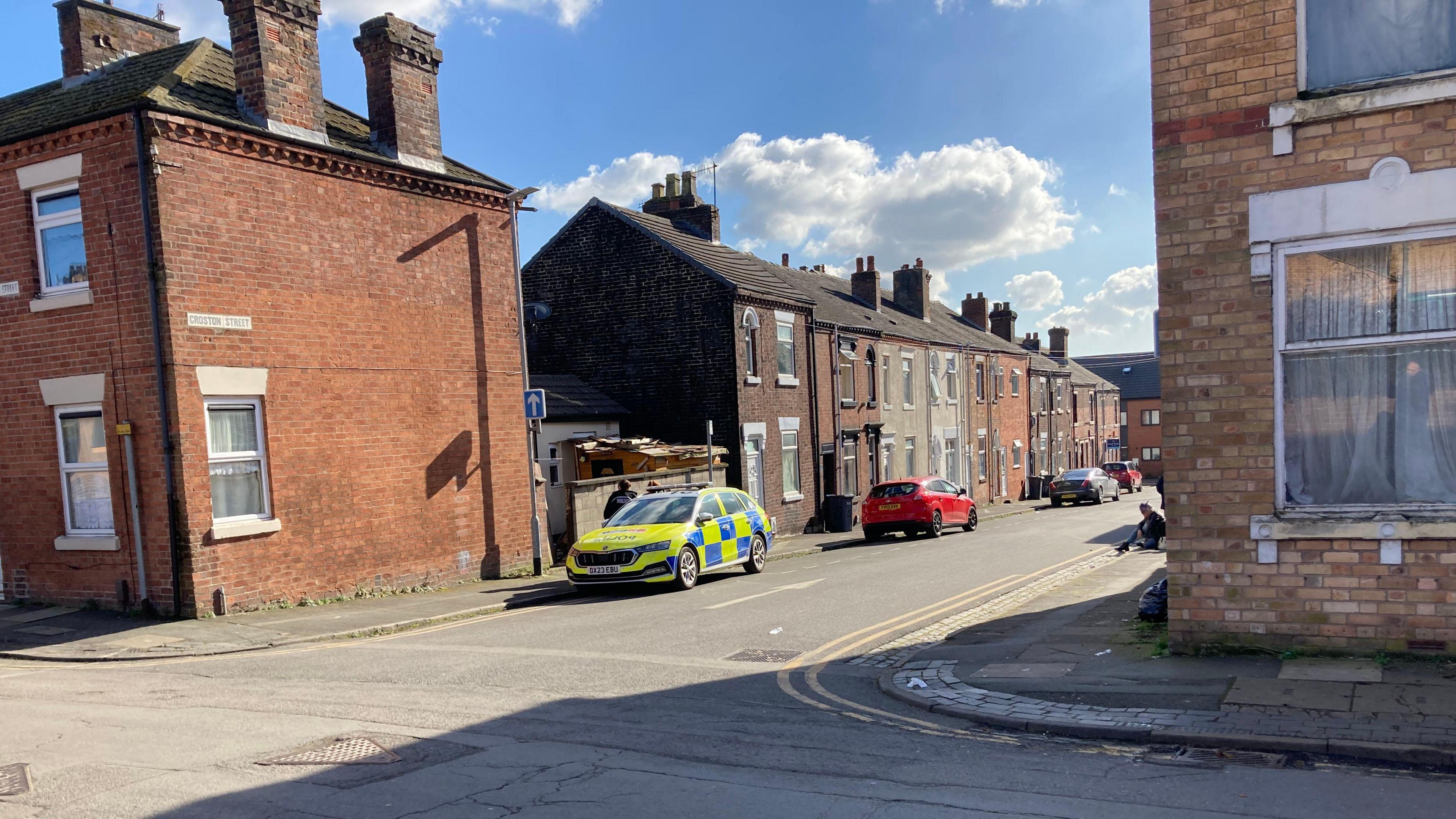 A residential street viewed from the entrance of a t-junction. A police car is parked next to a house on the left with two officers standing behind it.