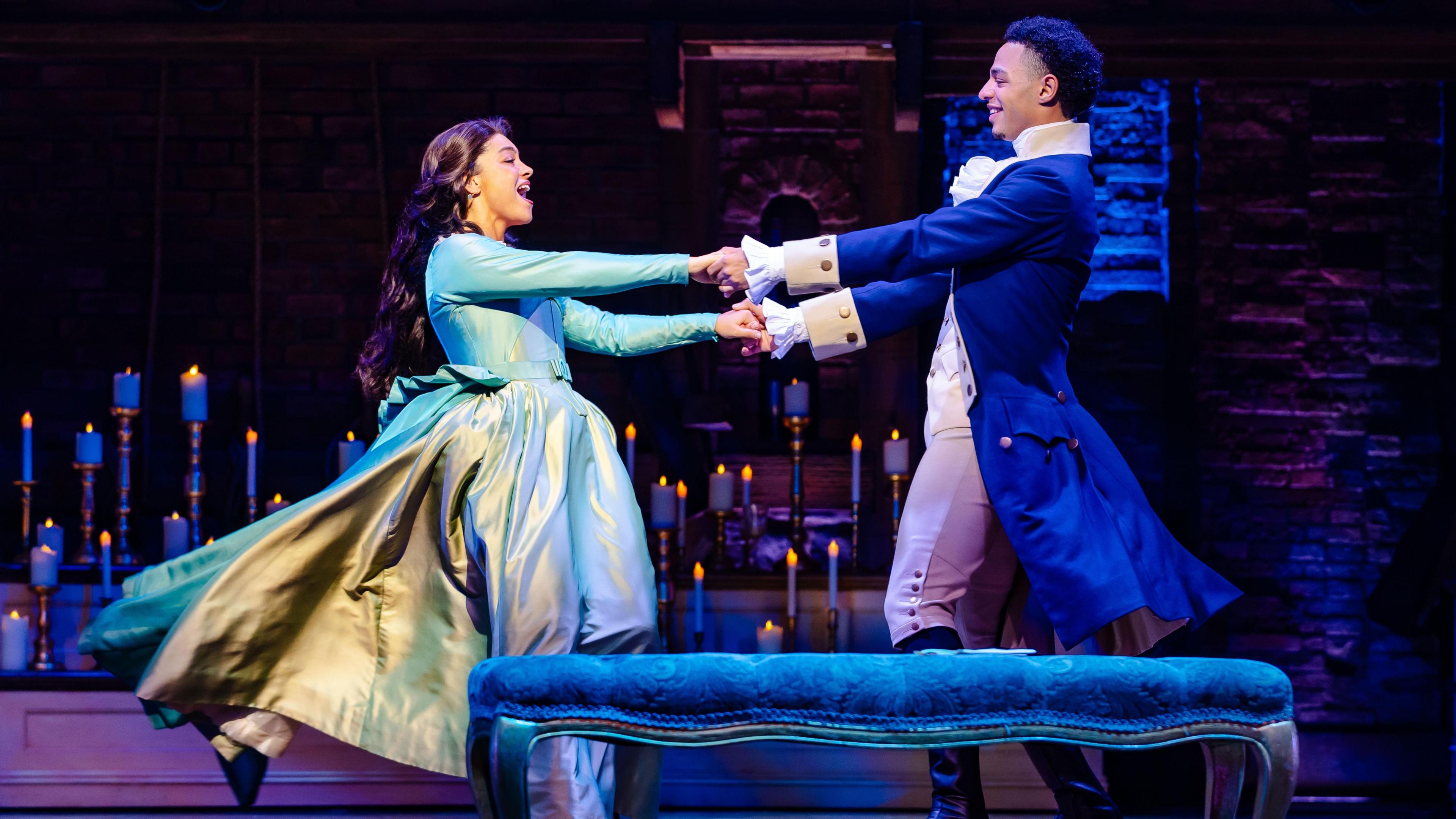 Marley Fenton, in costume in a long blue coat with a white cravat, dances with a woman on stage