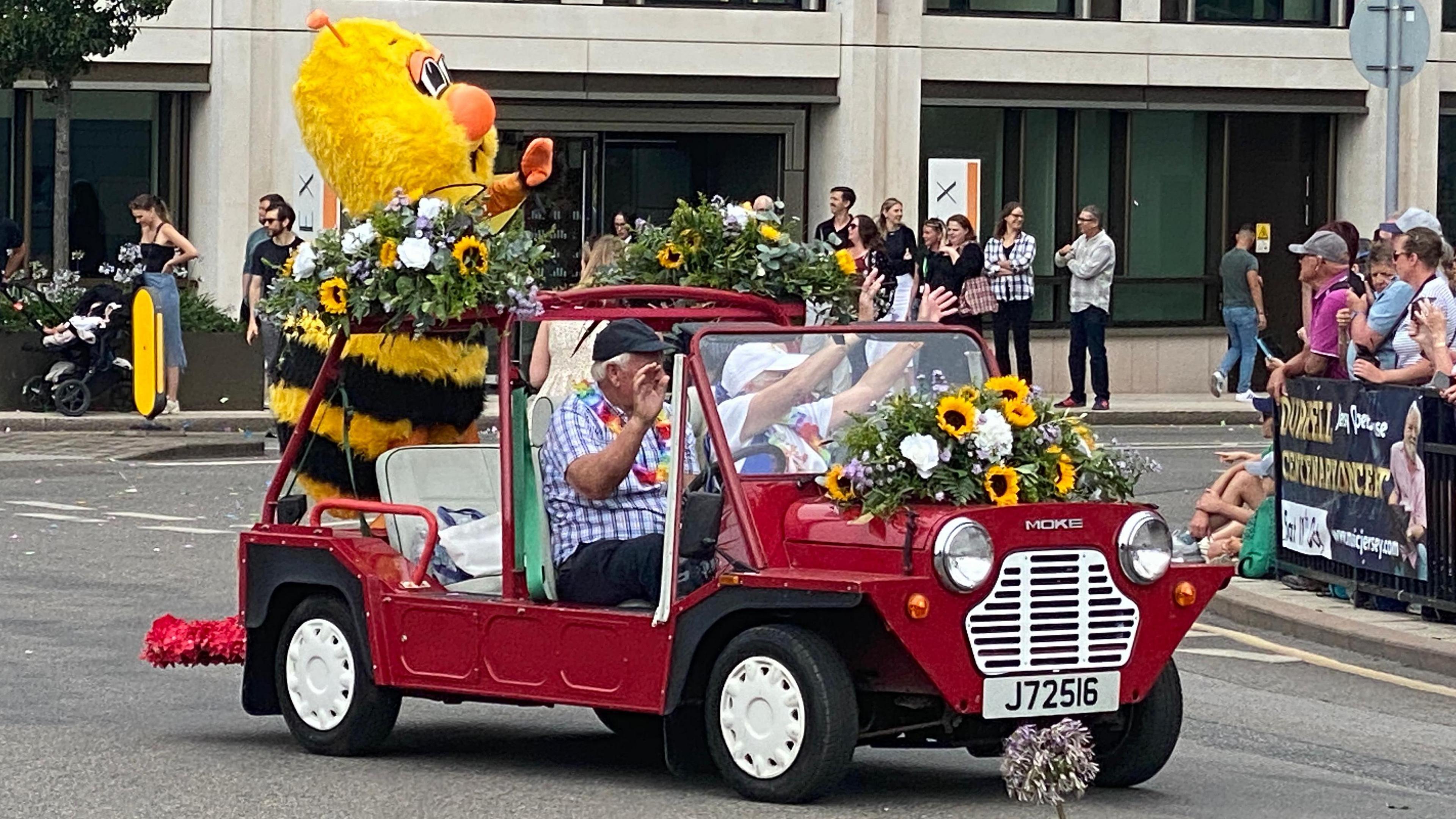A small open top car which has a giant black and yellow striped bee in the boot area. The car is decorated with flower arrangements featuring sunflowers. The driver and passenger are wearing flowers around their necks and are waving to the spectators.