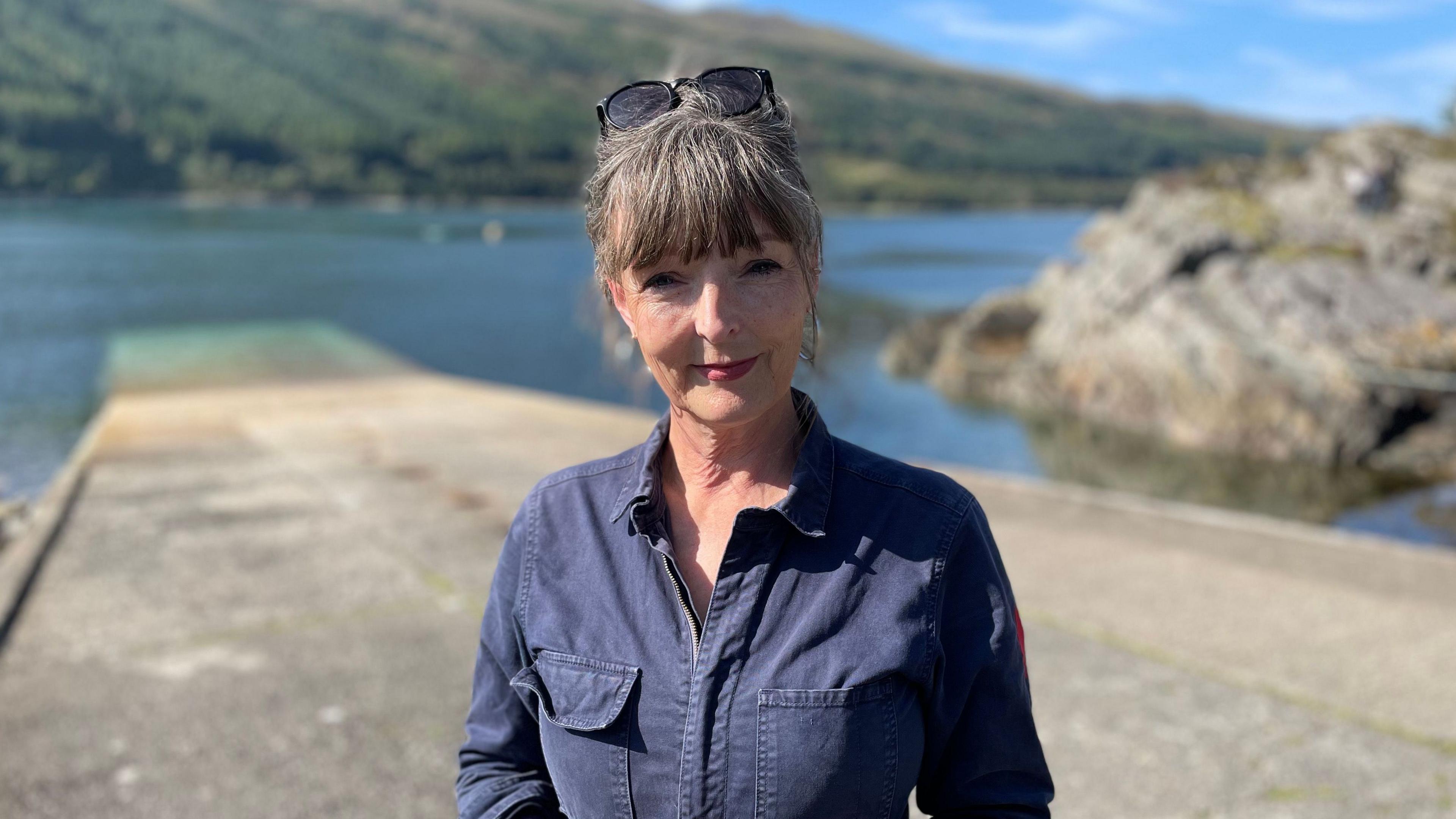 Boireannach le falt dorcha / liath.  Tha lèine ghorm dhorcha oirre agus tha speuclairean grèine air a ceann.  Tha i na seasamh air lamraig agus chithear caolas air a cùlaibh le sliabh le craobhan-giuthais air an taobh thall.  