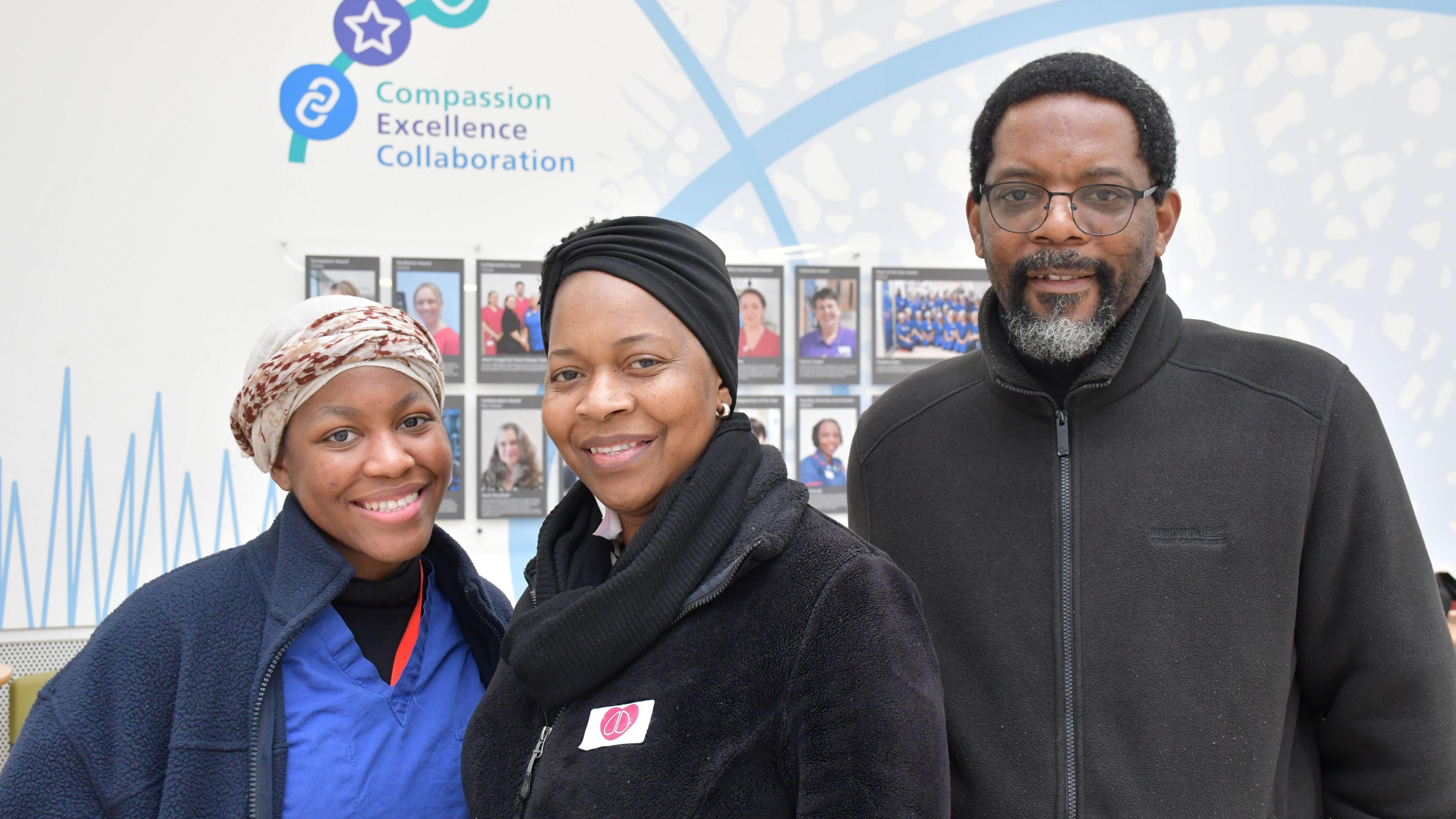 Ms Mushonga's daughter is smiling at the camera while standing next to her mum on the left. She has brown eyes and is wearing a blue fleece. Ms Mushonga's husband is standing to her right. He is wearing glasses and has black hair and black and grey facial hair.