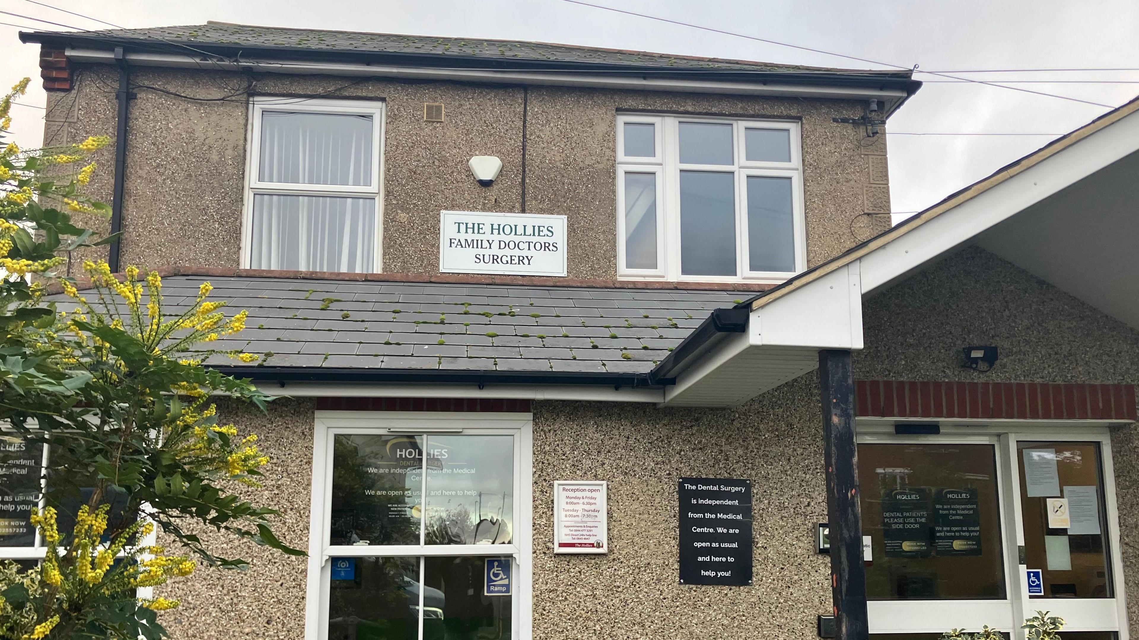 The front of The Hollies Surgery, which is a beige-coloured two-storey pebble-dash house. A sign reads THE HOLLIES FAMILY DOCTORS SURGERY.