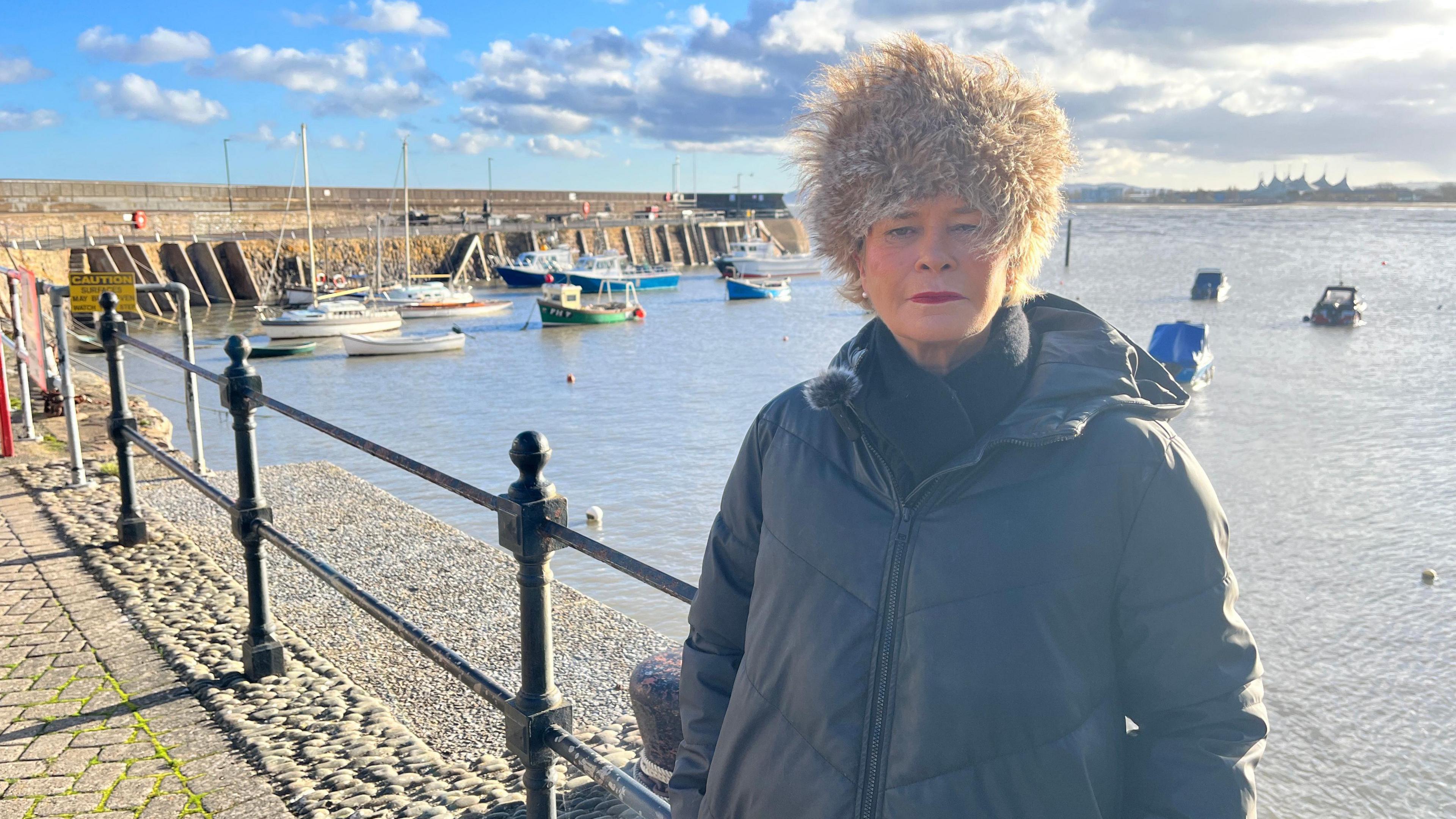 Rachel Gilmour is standing just to the right of the frame. She's wearing a Russian-style light brown hat, and a black puffer jacket. Behind her is Minehead harbour at high tide with boats bobbing on the water. In the distance, you can see the Butlins tent.