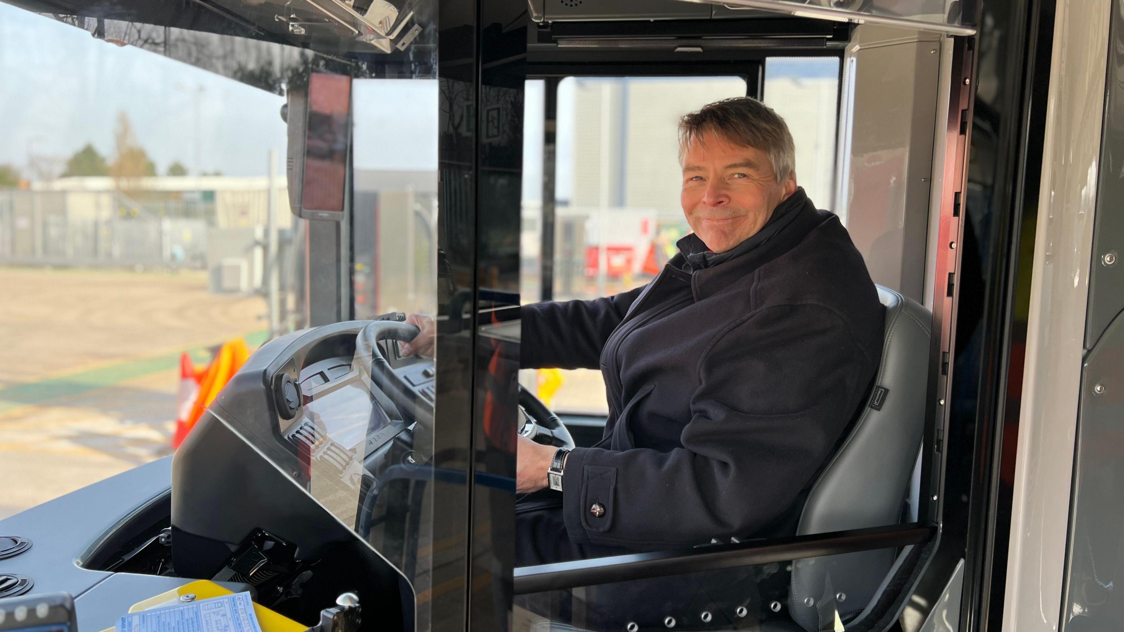 Councillor Andrew Gant sits in the driver's seat of a Stagecoach bus, he's smiling at the camera with his hands on the steering wheel. He has short grey hair and wears a black collared wool coat.