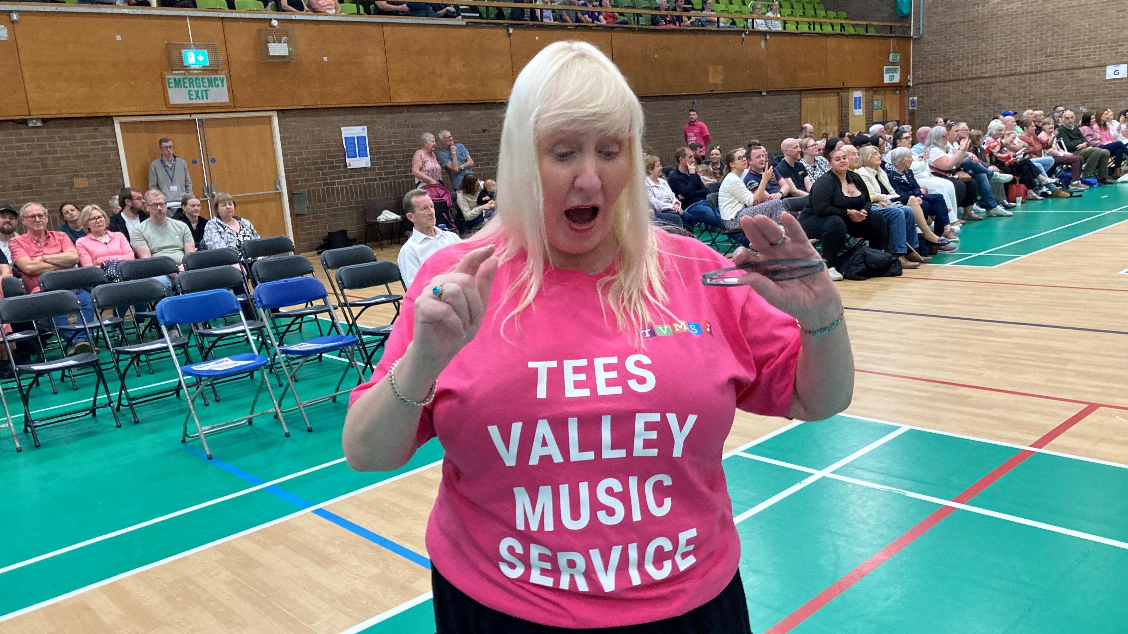 Susan Robertson, in a pink t-shirt reading Tees Valley Music Service, singing with parents watching in the background.