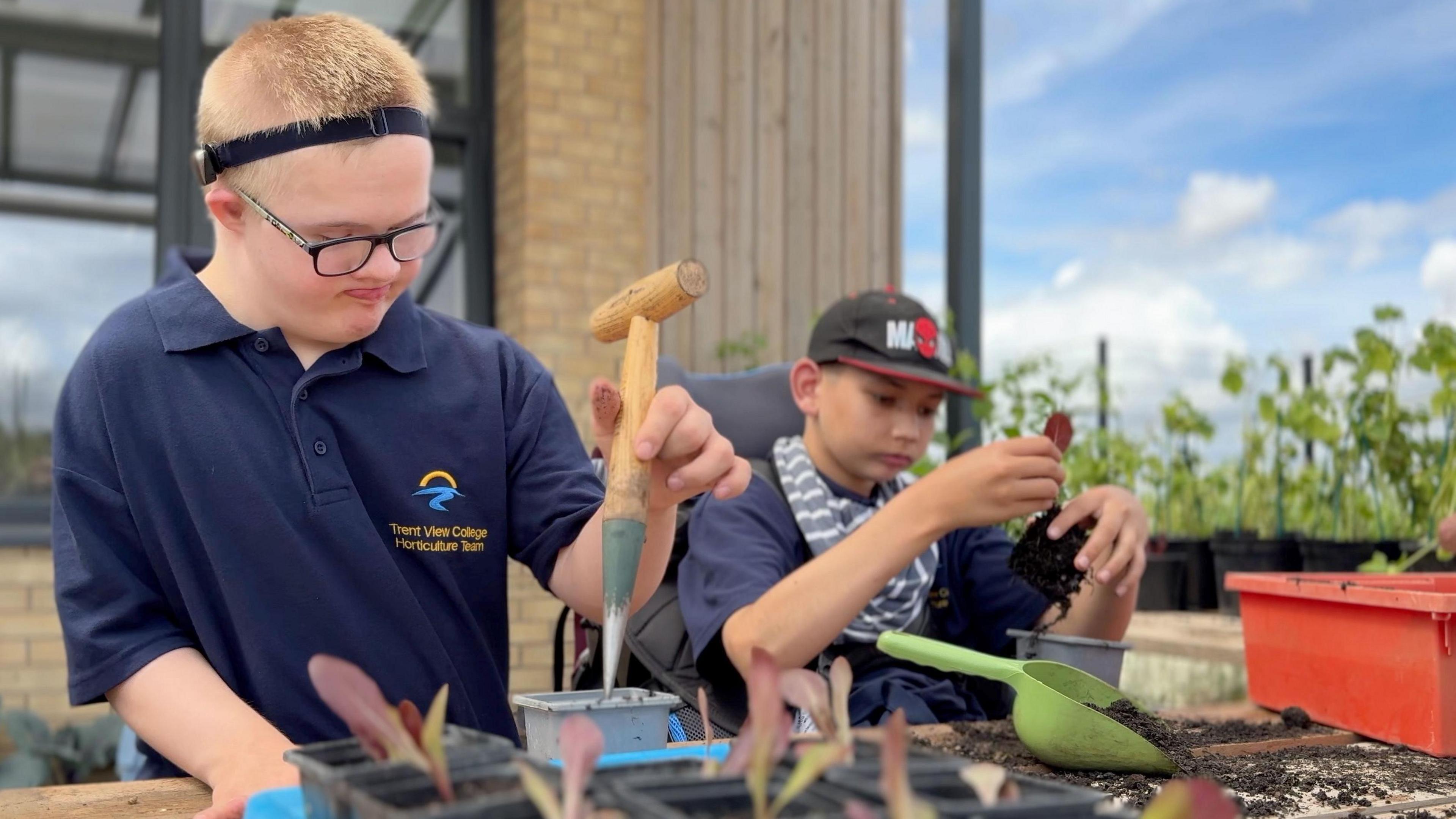 Two young men both wearing navy blue school uniforms use tools and pots. Lewis has blonde hair and is working with a grey plastic plant pot on the left. Blake has a green trowel and is examining a red leaf.