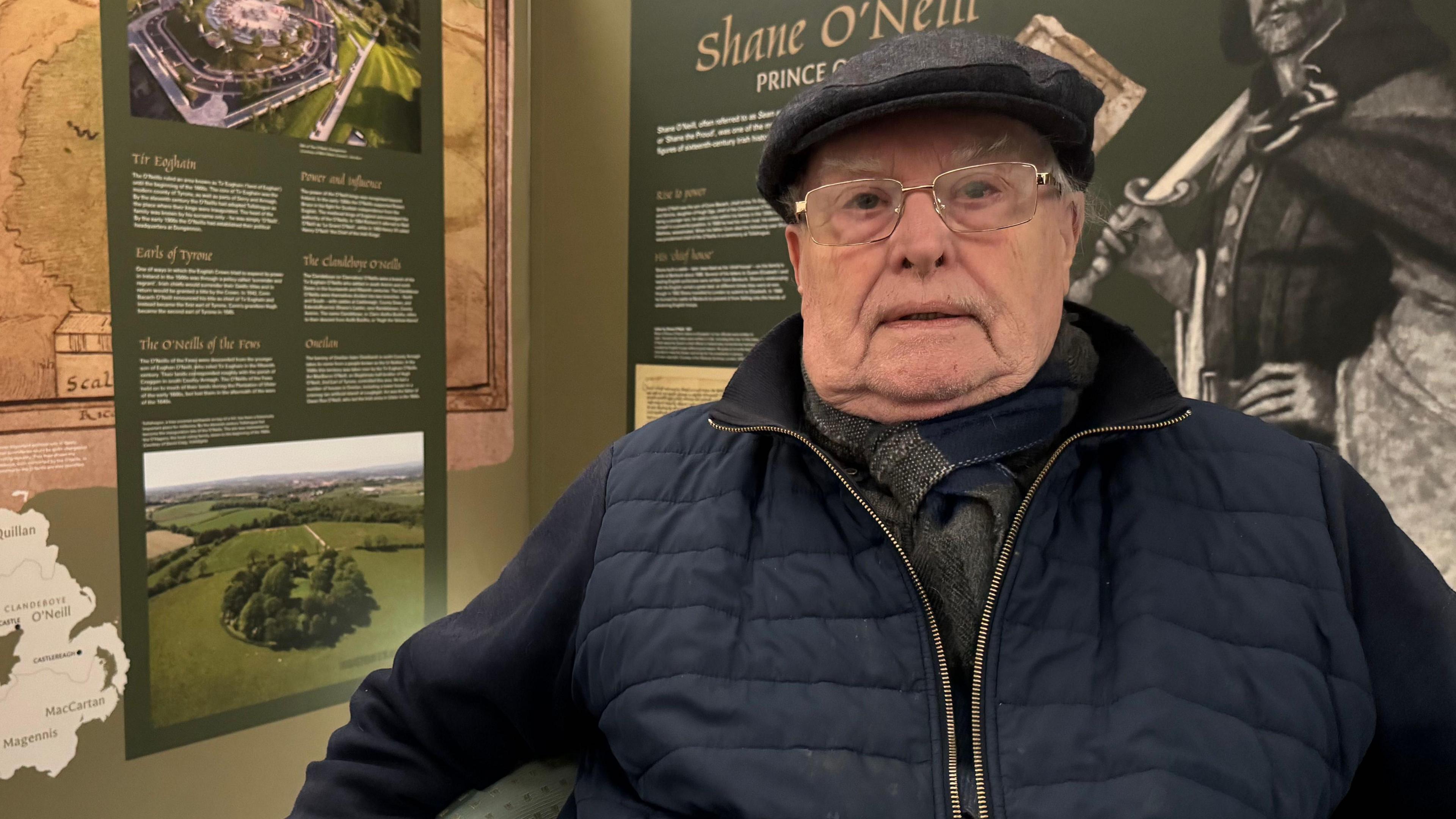 An older man is sitting in a corner looking into the camera. He is wearing metal rim glasses, a dark cap and scarf, and a navy coat. Behind him are information panels on the history of Shane O'Neill. 