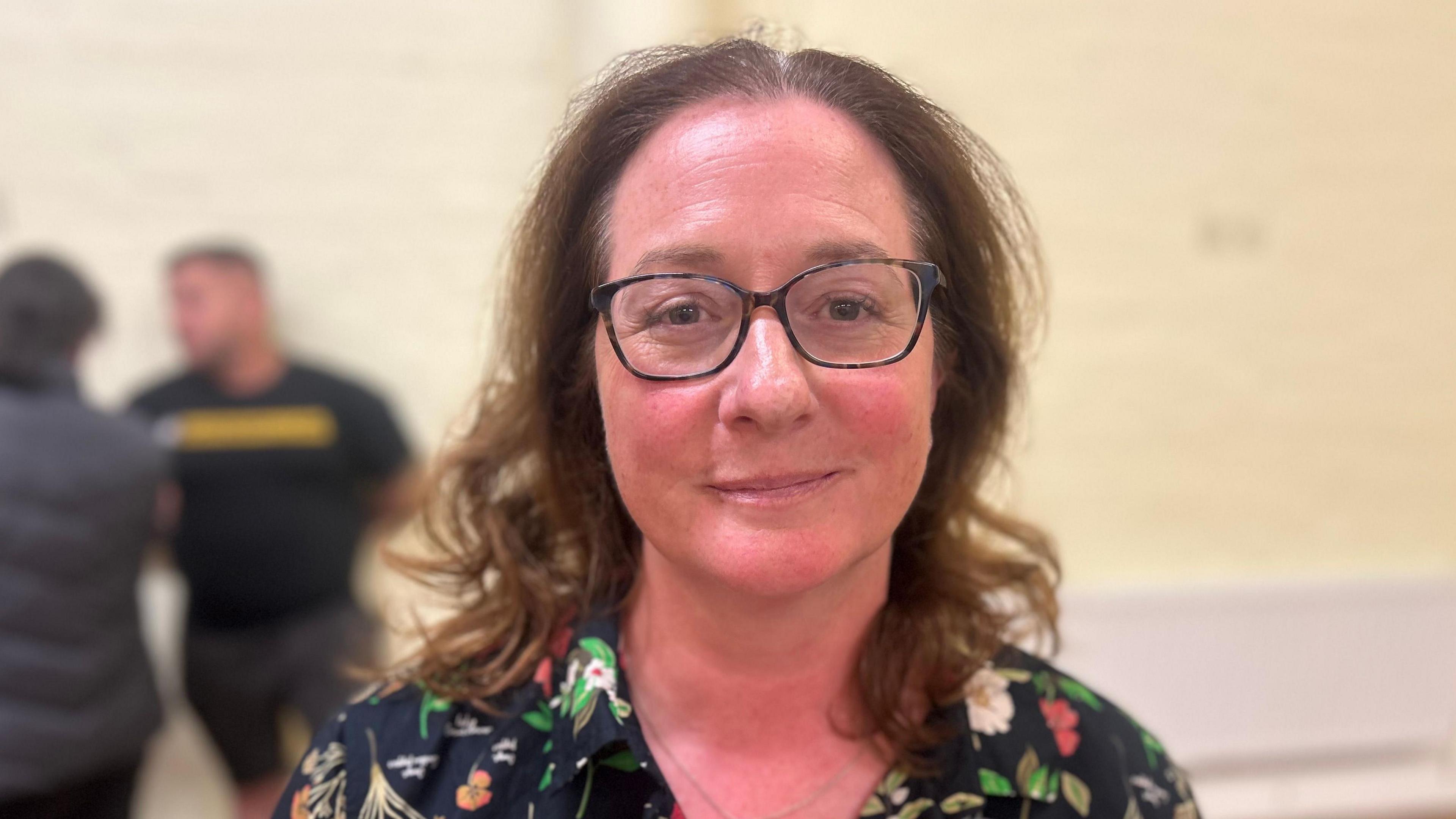 Vicki Wade looking at the camera. She has brown curly hair and glasses and is wearing a flowery shirt.