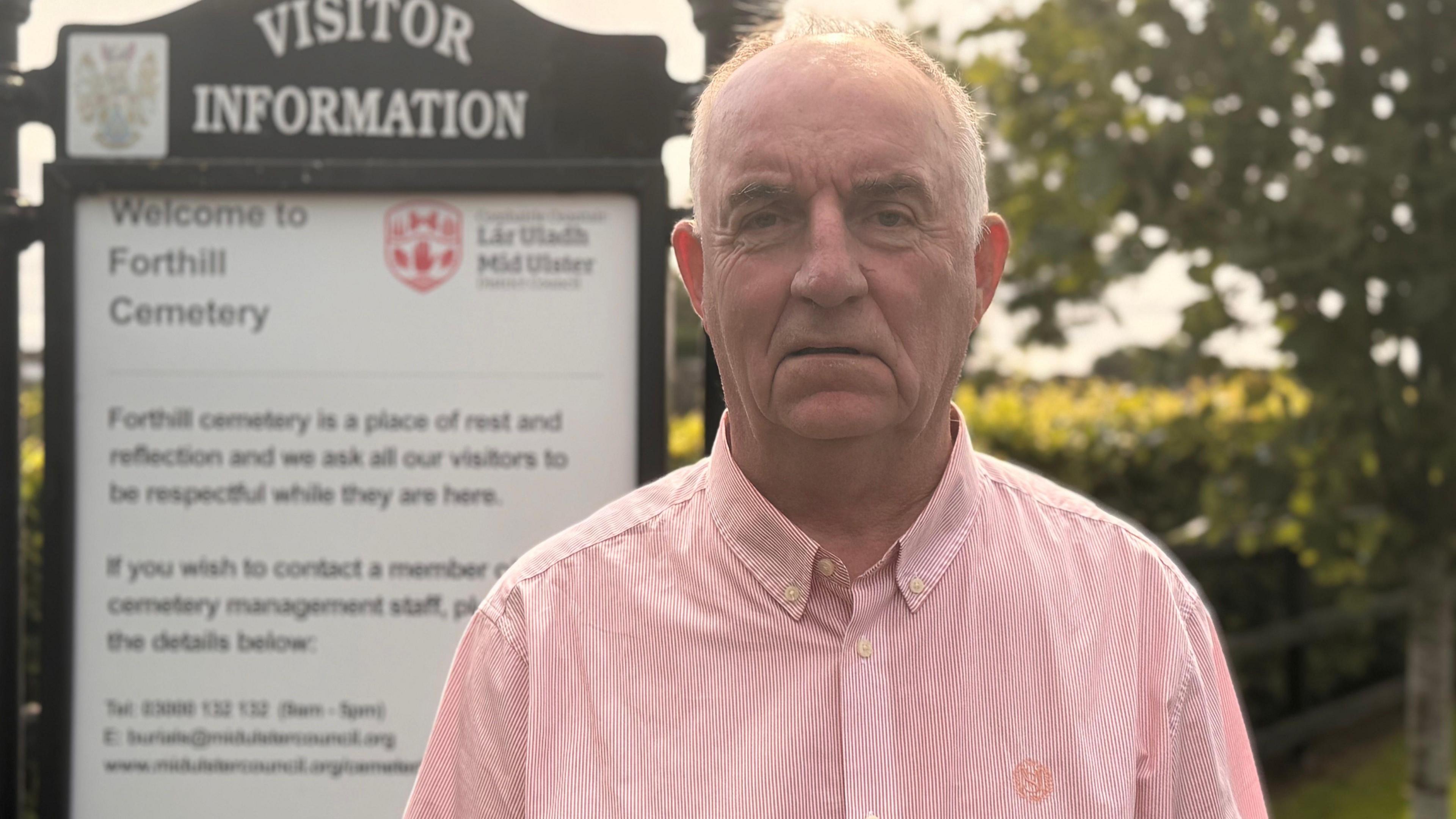 Trevor Wilson stands in front of a cemetery's information board. He is wearing a pink shirt and has a stern serious expression on his face. He is balding but has grey hair. The sign is behind his right shoulder, and reads welcome to forthill cemetery. A tree is visible behind his left shoulder.