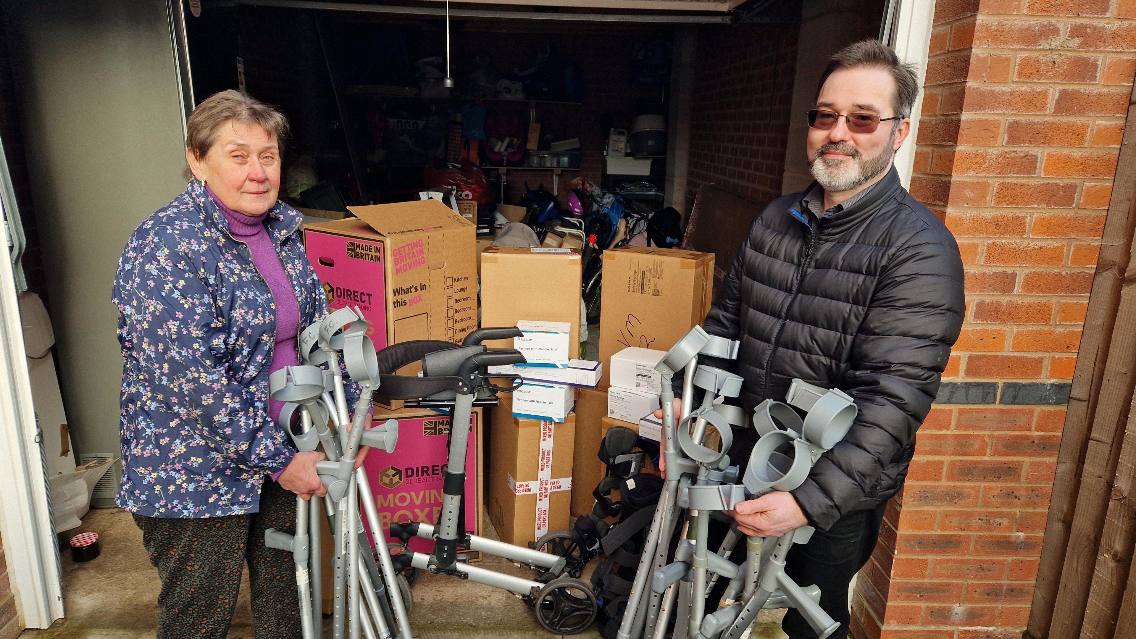 A woman with short dark hair in a blue patterned jacket, holding a selection of crutches, stood next to a man with dark hair and a grey beard, wearing a black jacket and also holding a collection of crutches