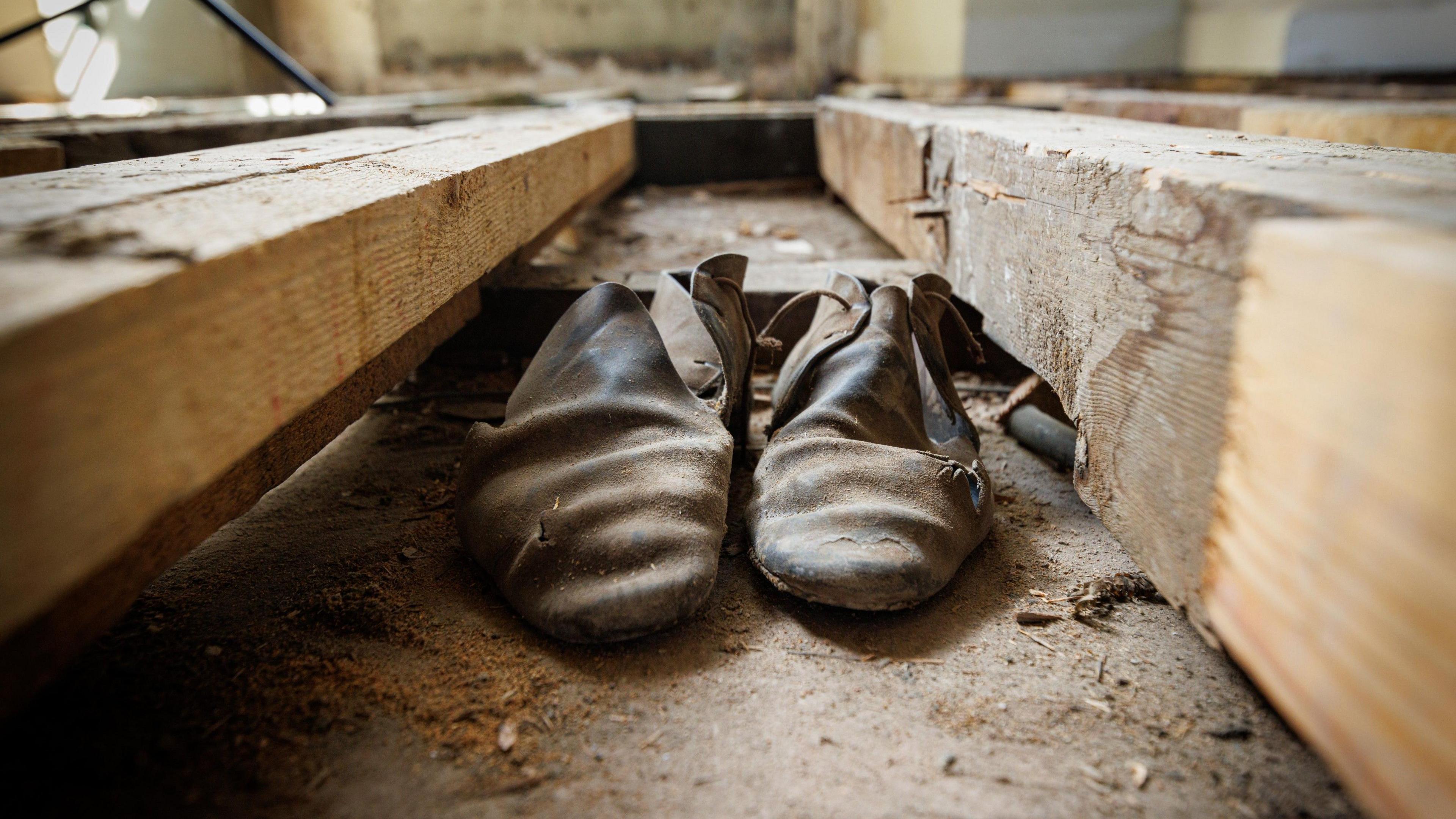 A very worn and dusty pair of black men's shoes sits between two floorboards.