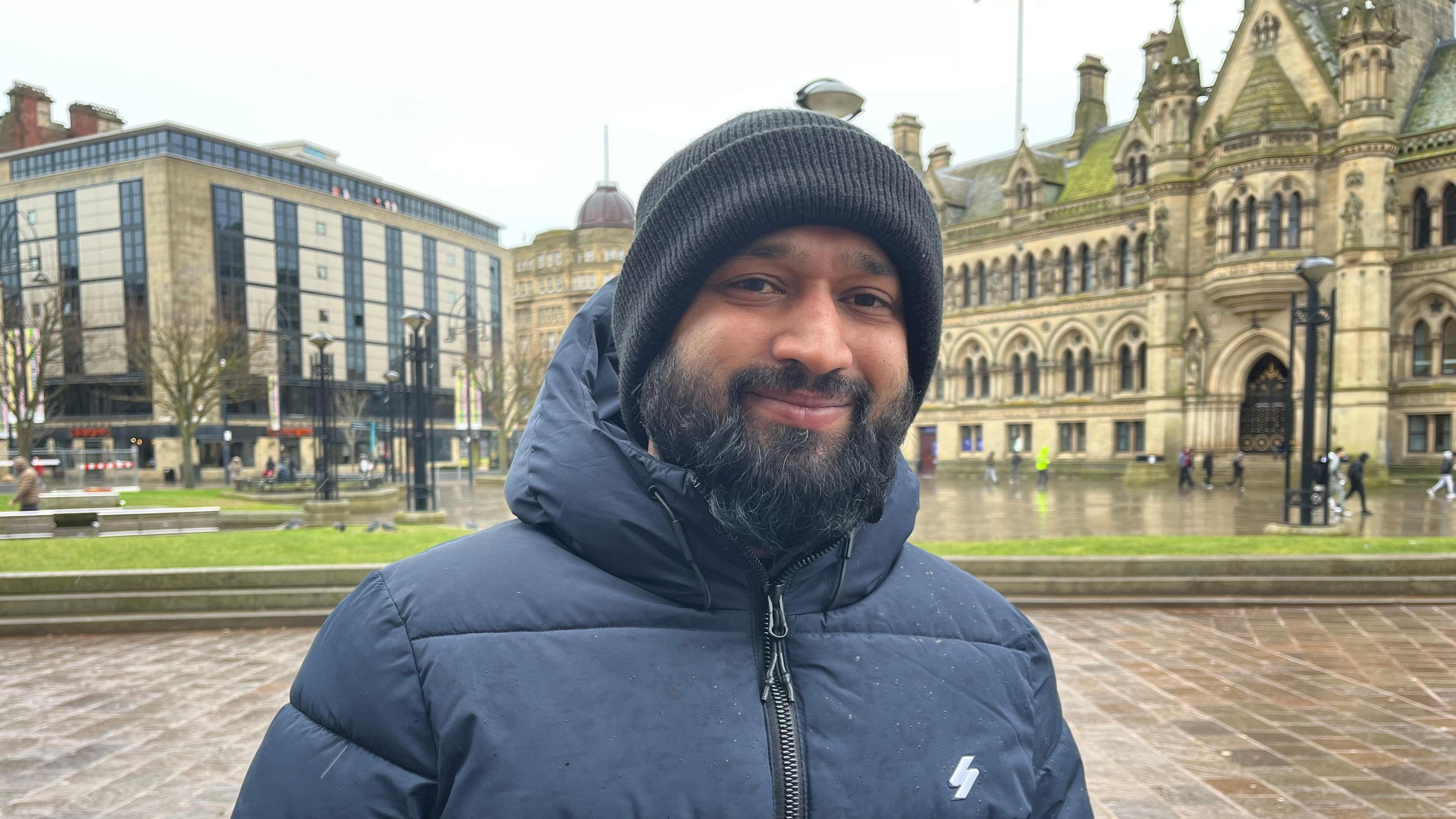 A man with a beard in a blue puffer coat and black hat is standing in front of a Victorian building